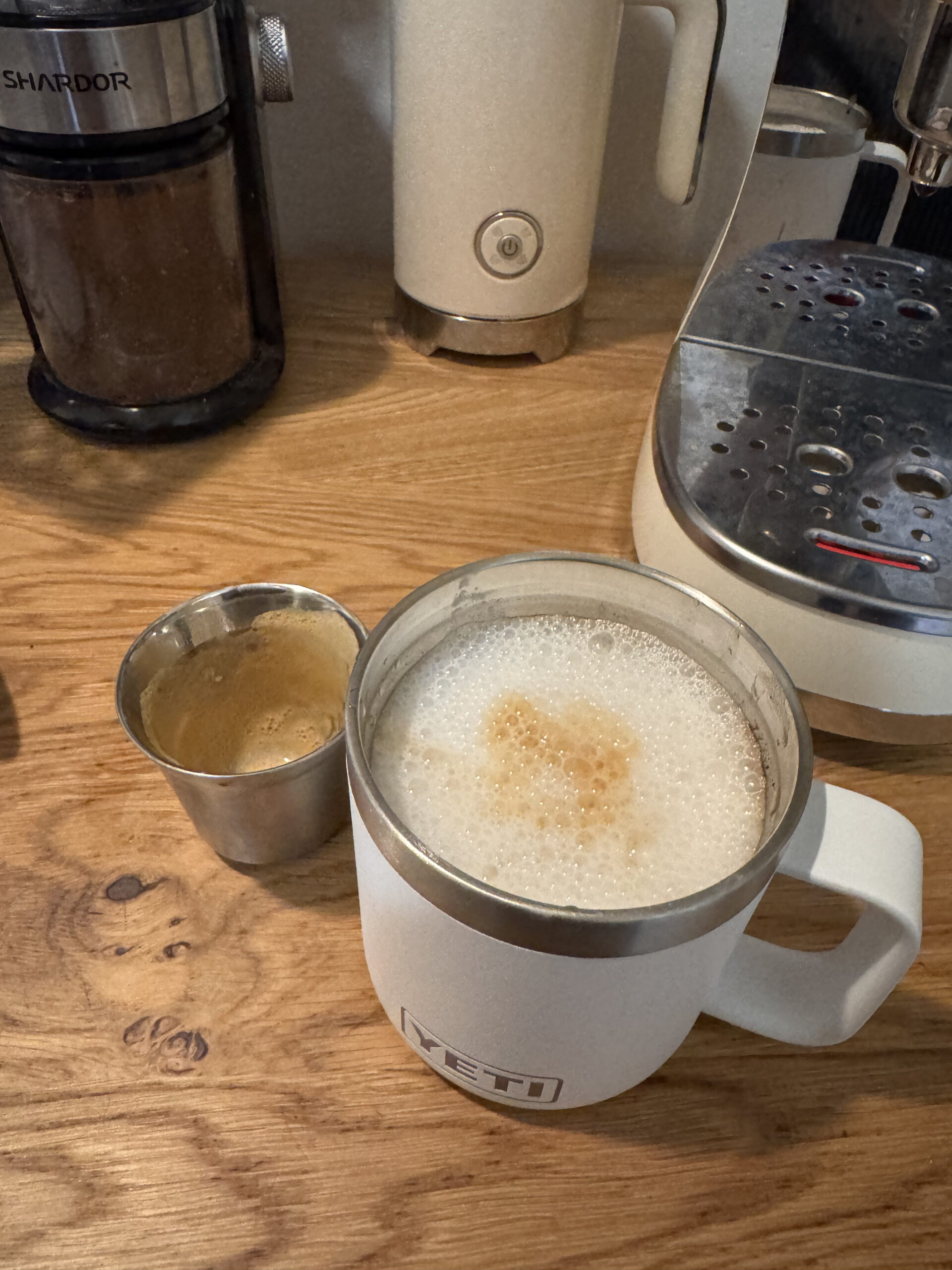 A mug of frothy coffee sits on a wooden table next to a small metal cup of espresso. Coffee maker and grinder are in the background.