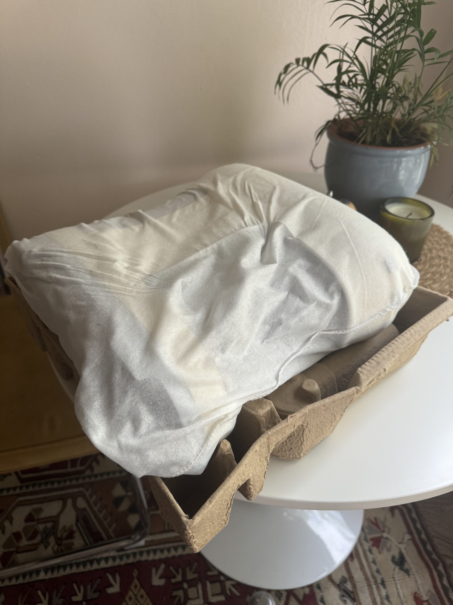 A white pillow covered with a case is resting on a cardboard tray on a white table next to a potted plant and a candle.