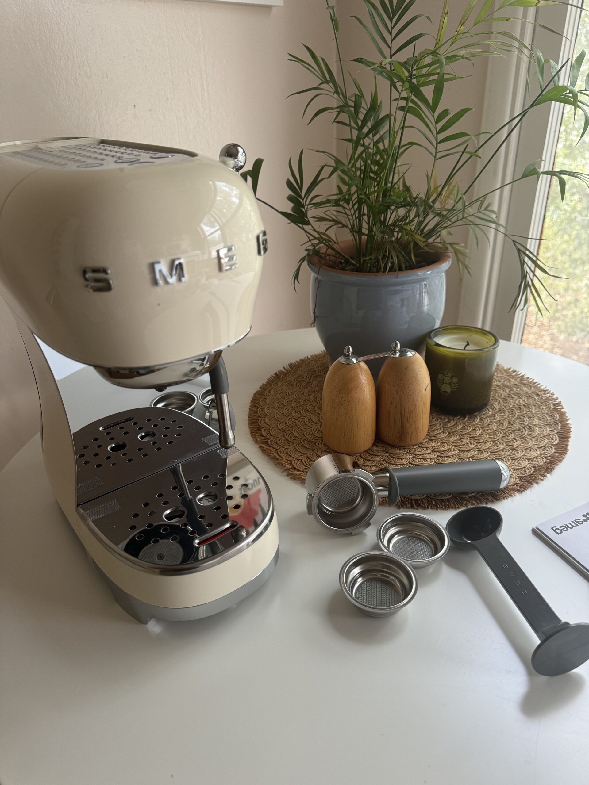 A Smeg espresso machine on a round table with coffee accessories, a plant, a candle, and wooden salt and pepper shakers nearby.