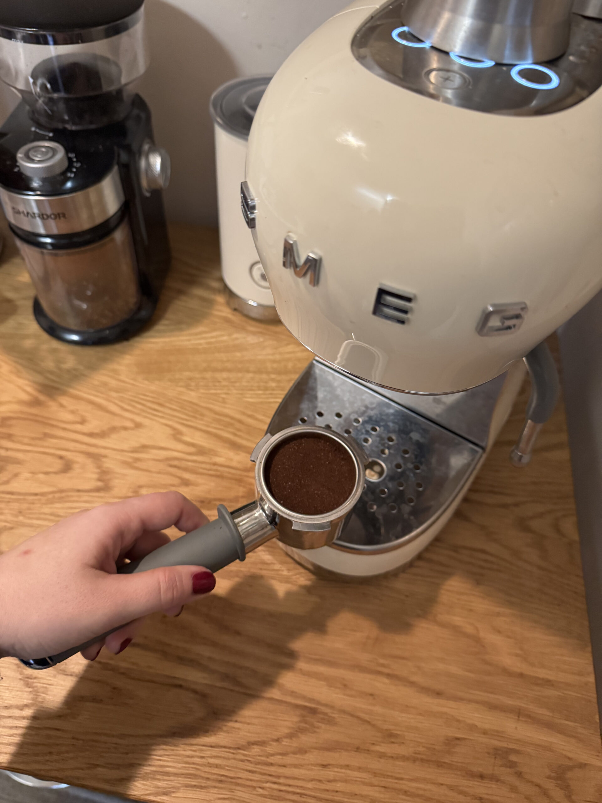 A hand holds a portafilter with coffee grounds in front of a cream-colored espresso machine on a wooden counter. A grinder and a sealed container are seen in the background.