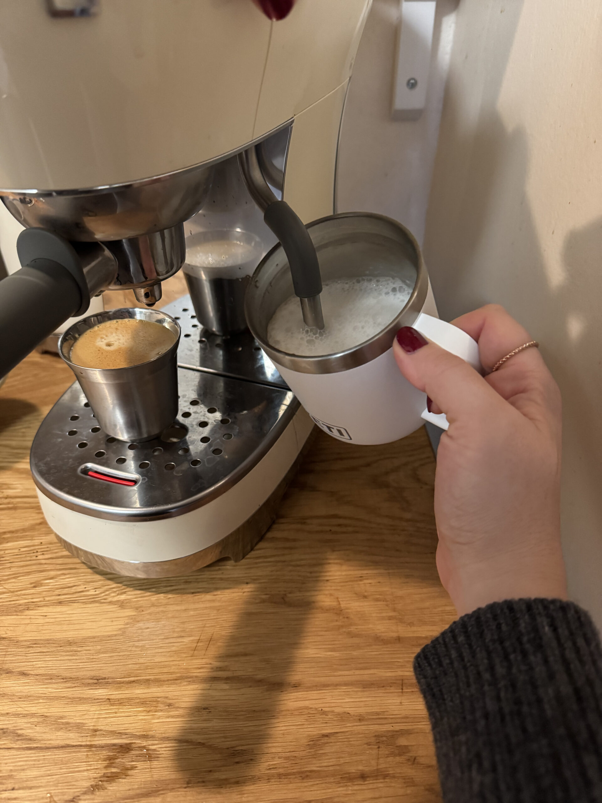 A person uses an espresso machine to steam milk in a metal cup while an espresso shot is being brewed next to it.