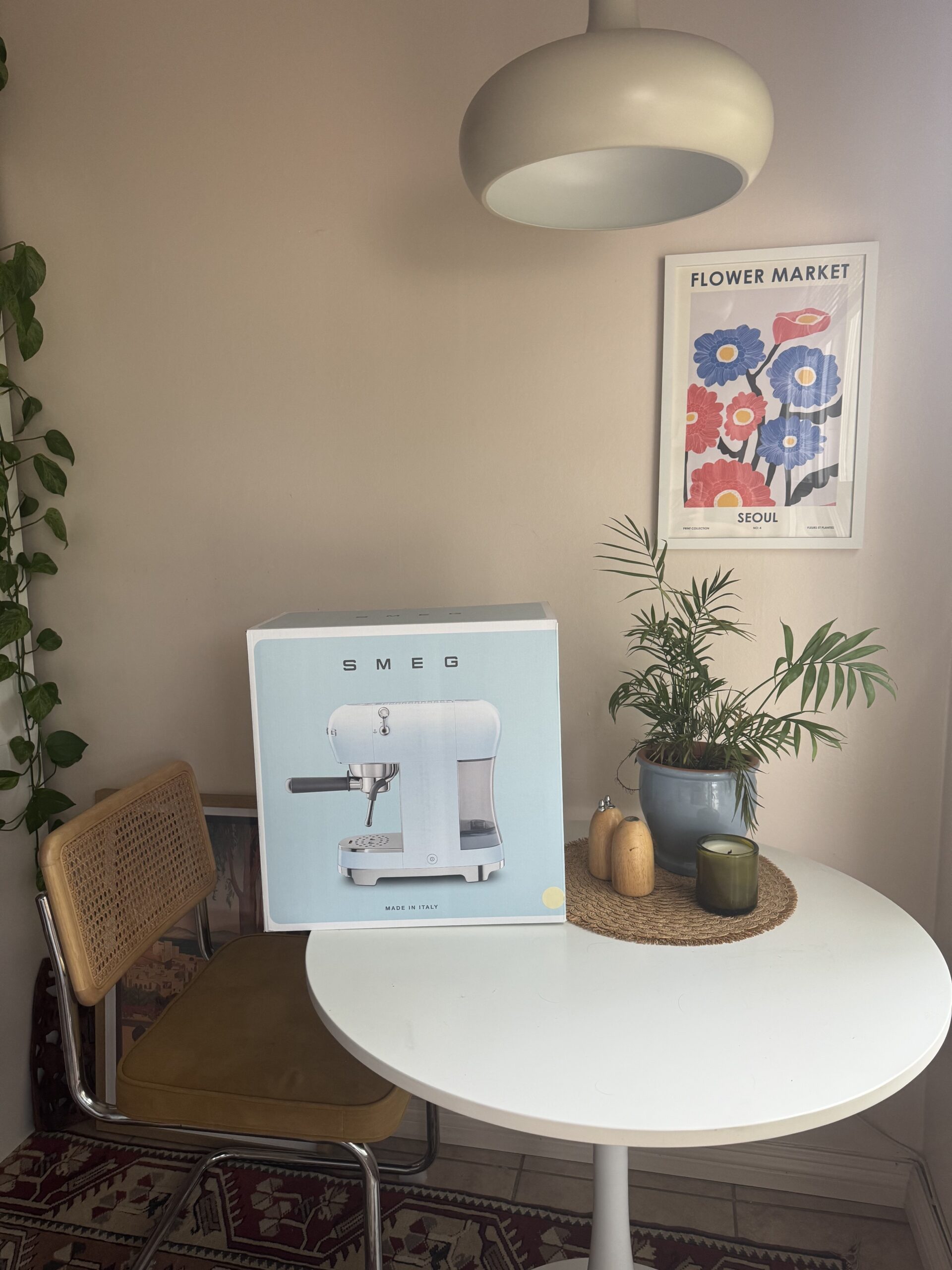 A boxed Smeg appliance sits on a round white table with plants and a candle. A chair is beside the table, and a "Flower Market" poster is on the wall.