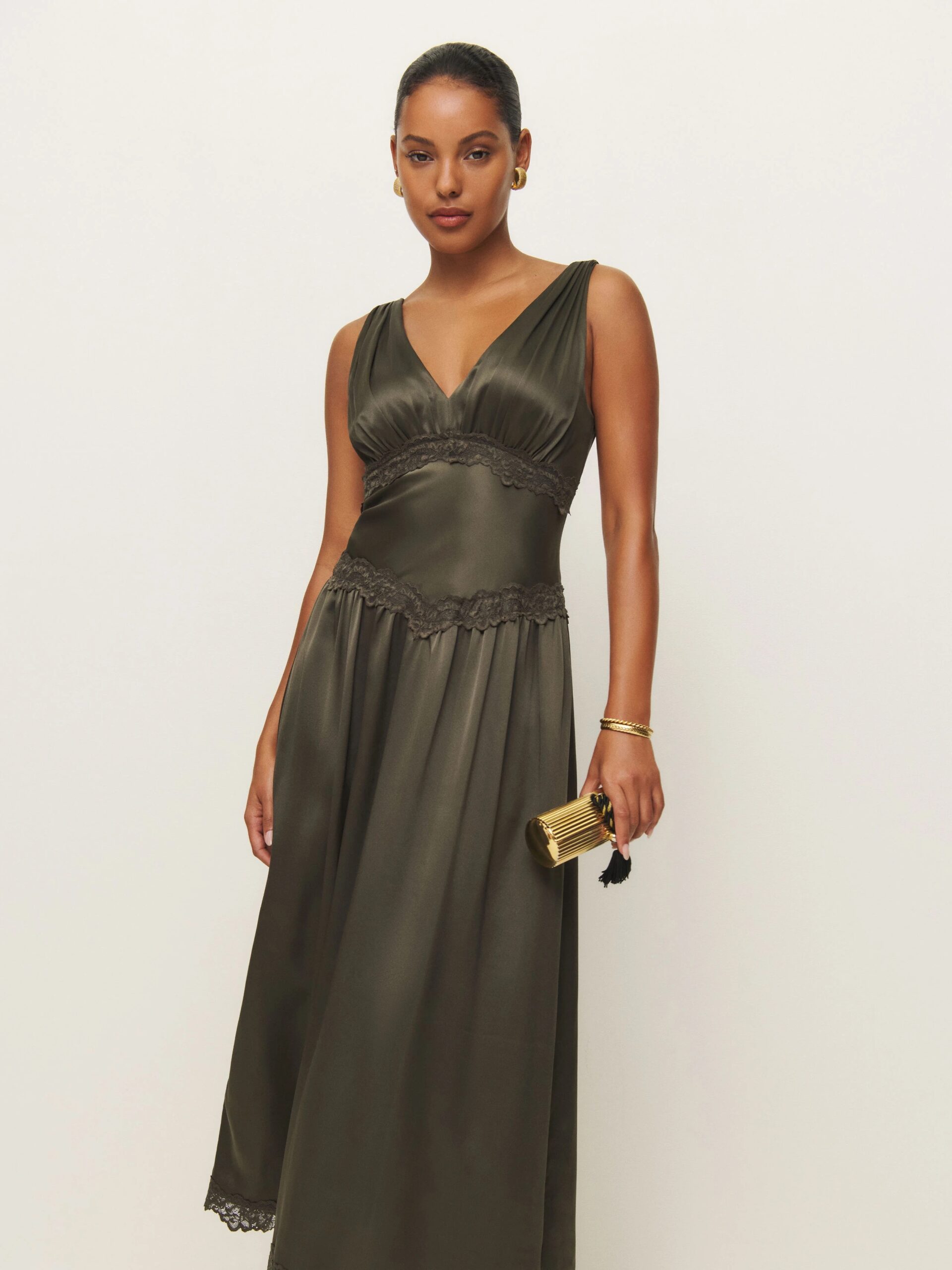 A person in a sleeveless, olive-green dress with lace details holds a small gold clutch.