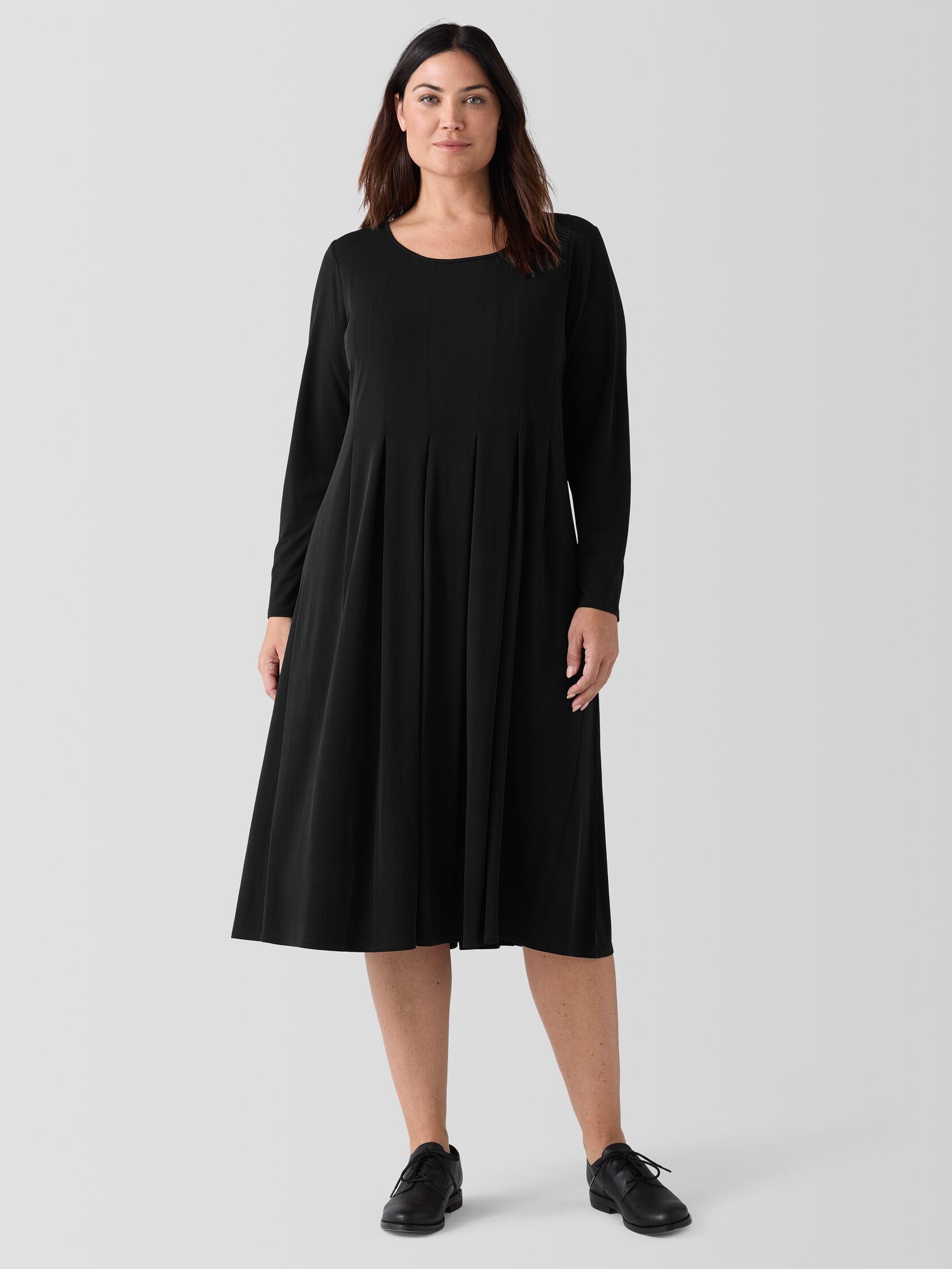 A woman wearing a long-sleeved, pleated black dress and black shoes stands against a plain background.