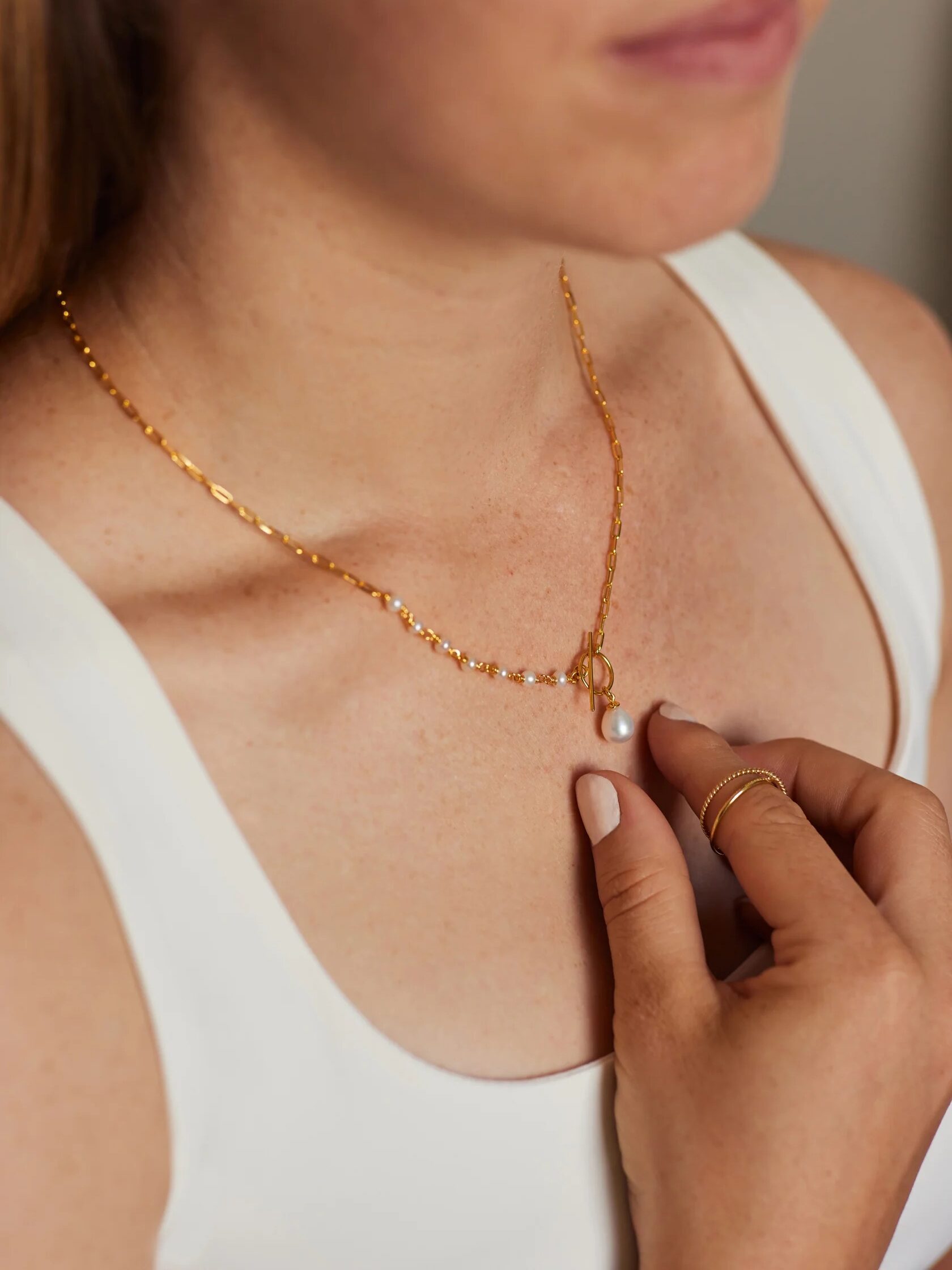 A person wearing a white tank top holds a gold necklace with a small pendant and pearls.