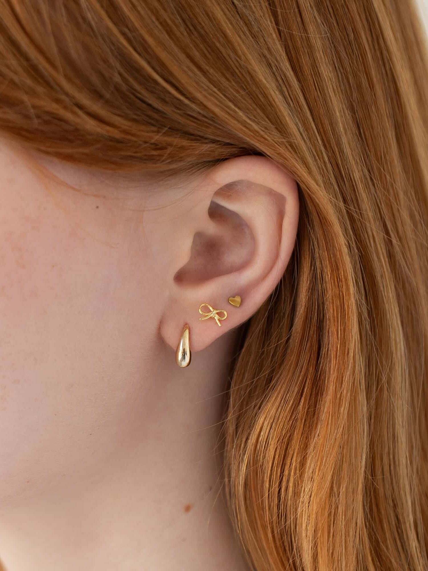 Close-up of a person's ear adorned with three gold earrings: a teardrop, a bow, and a heart. The person has long, reddish hair.