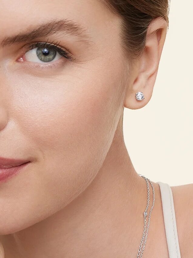 Woman wearing a diamond stud earring and a necklace, smiling slightly against a neutral background.