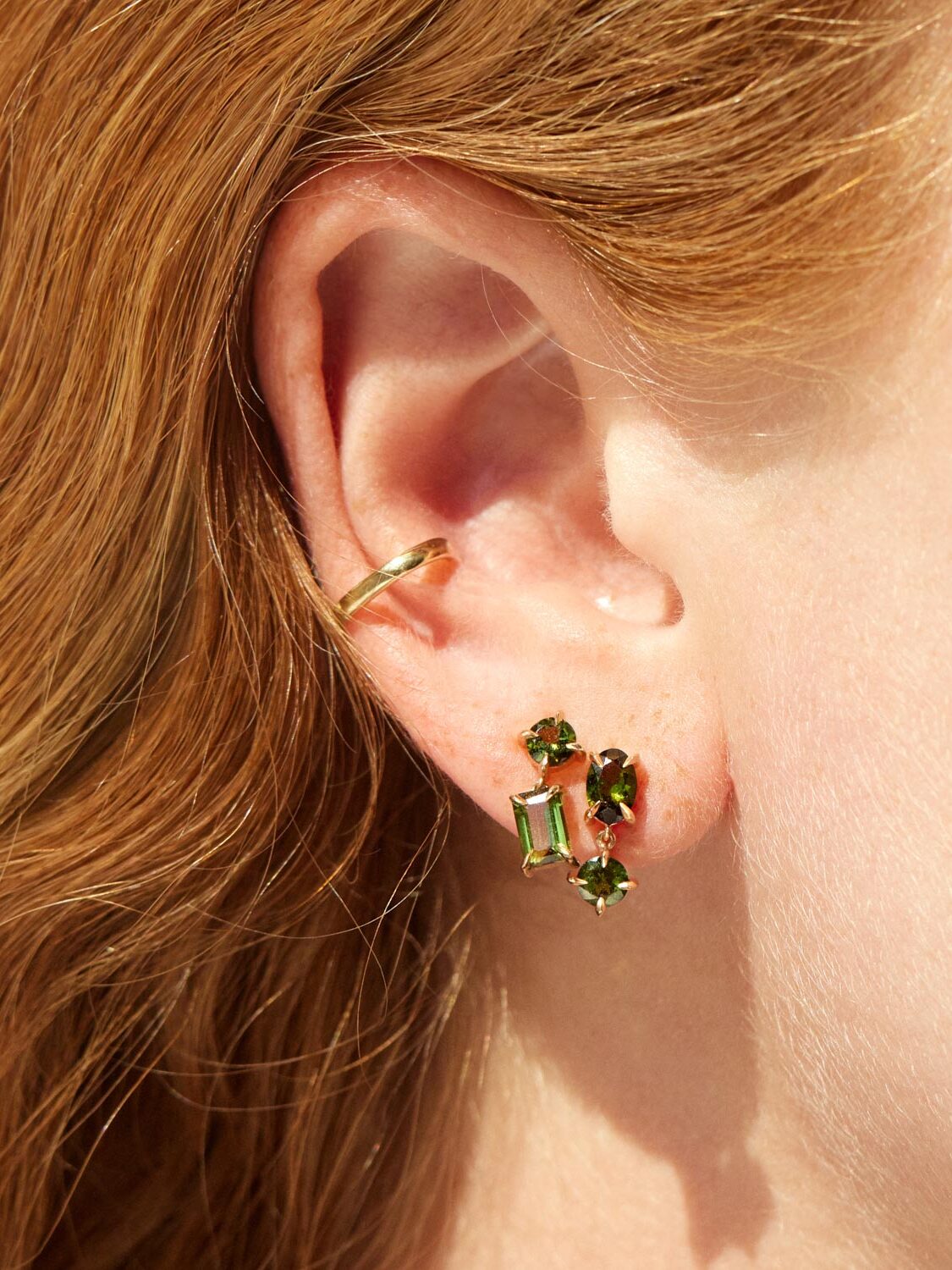 Close-up of an ear adorned with three green gemstone earrings and a gold ear cuff.