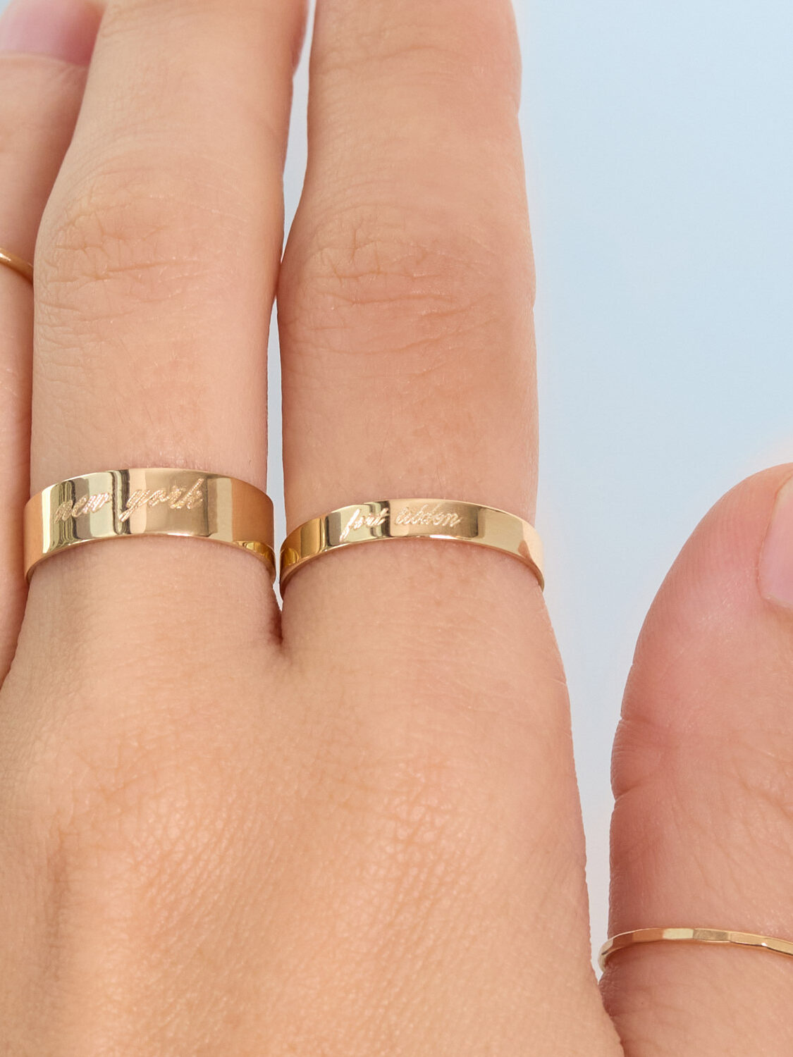 Two gold rings engraved with "New York" and "Boston" worn on adjacent fingers on a hand with a light skin tone.