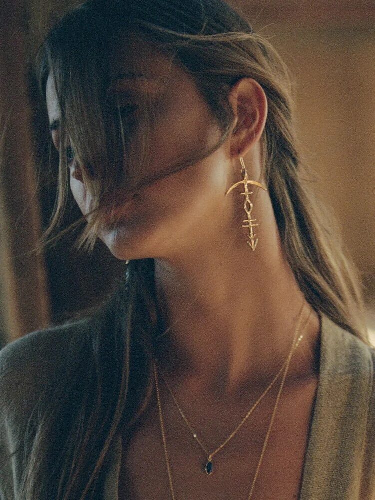 A person with long hair partially covering their face is wearing layered necklaces and a long, gold earring, standing in a softly lit room.