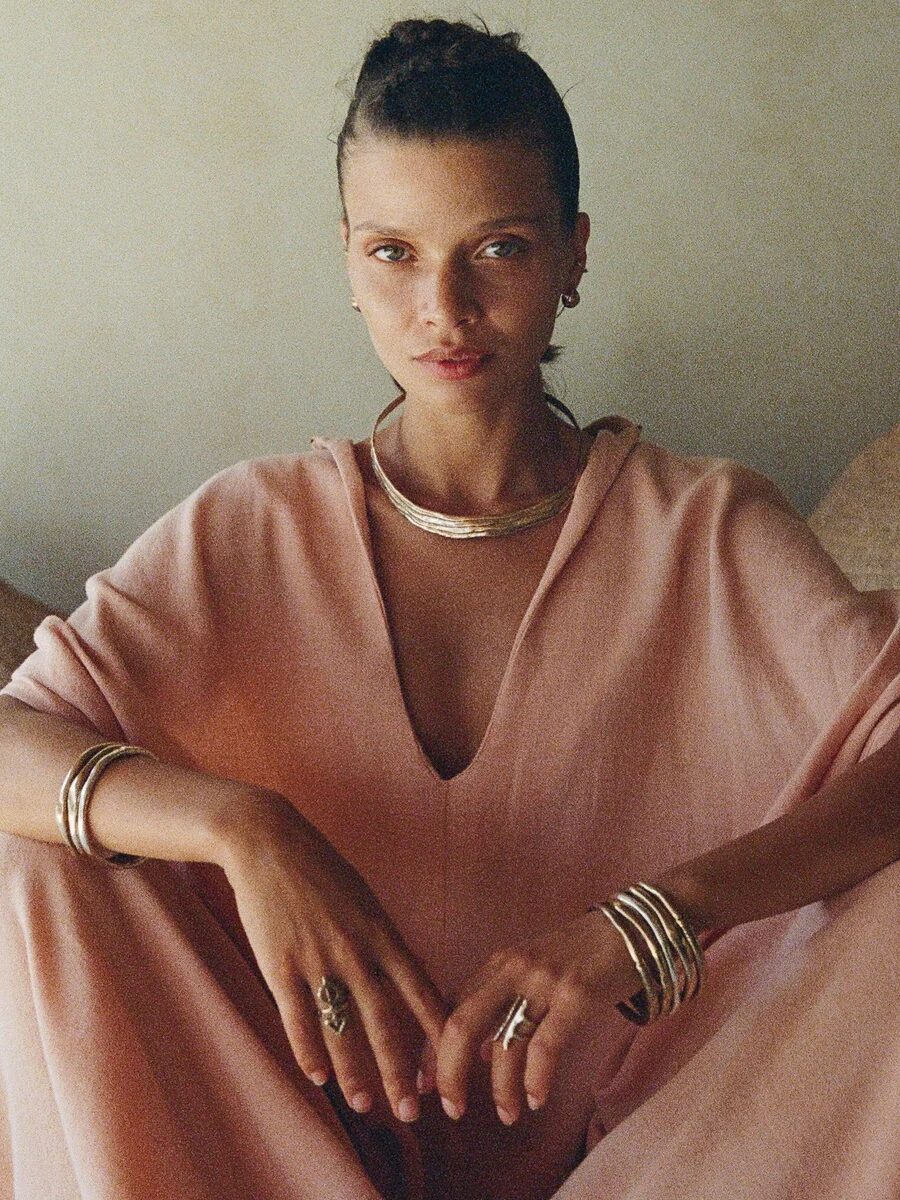 Woman in a pink robe sits cross-legged, wearing gold jewelry. Baskets are visible in the background.