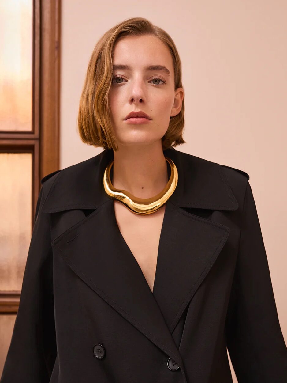 A person with short hair wears a black coat and a chunky gold necklace, standing indoors near a wooden-framed window.