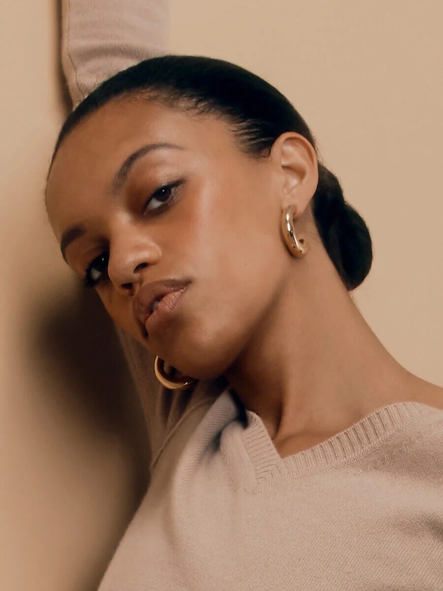 Person with earrings and a neutral expression poses with one arm raised against a beige background.