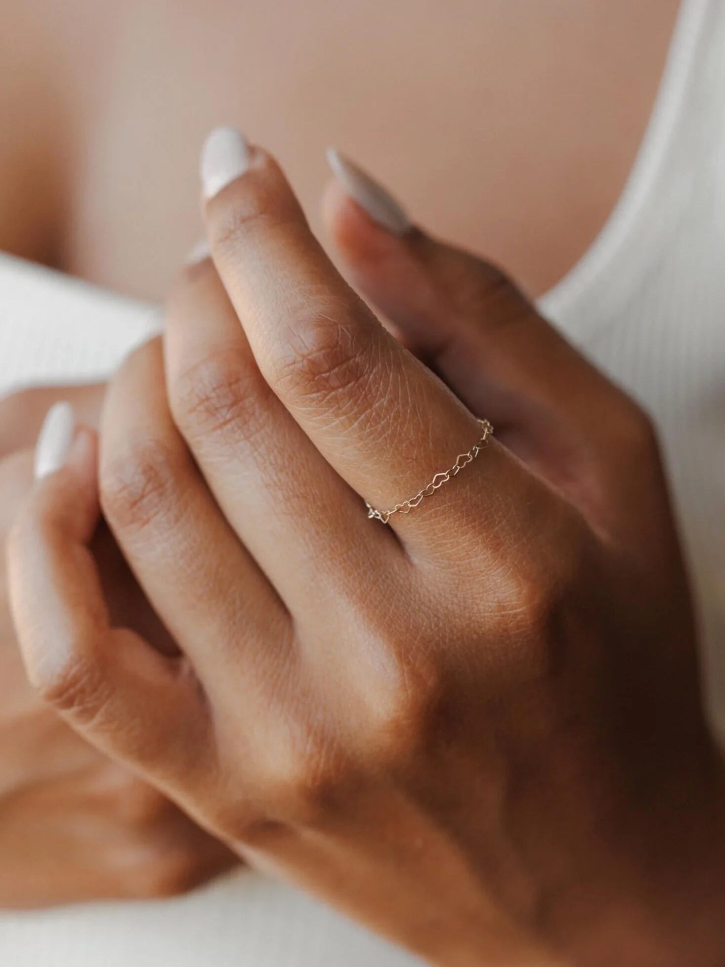 A hand with a delicate gold chain ring on the finger, showing neatly manicured nails against a white ribbed fabric background.