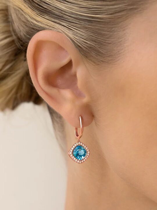 A person wearing a hoop earring with a blue gemstone and surrounding small diamonds, against a plain background.