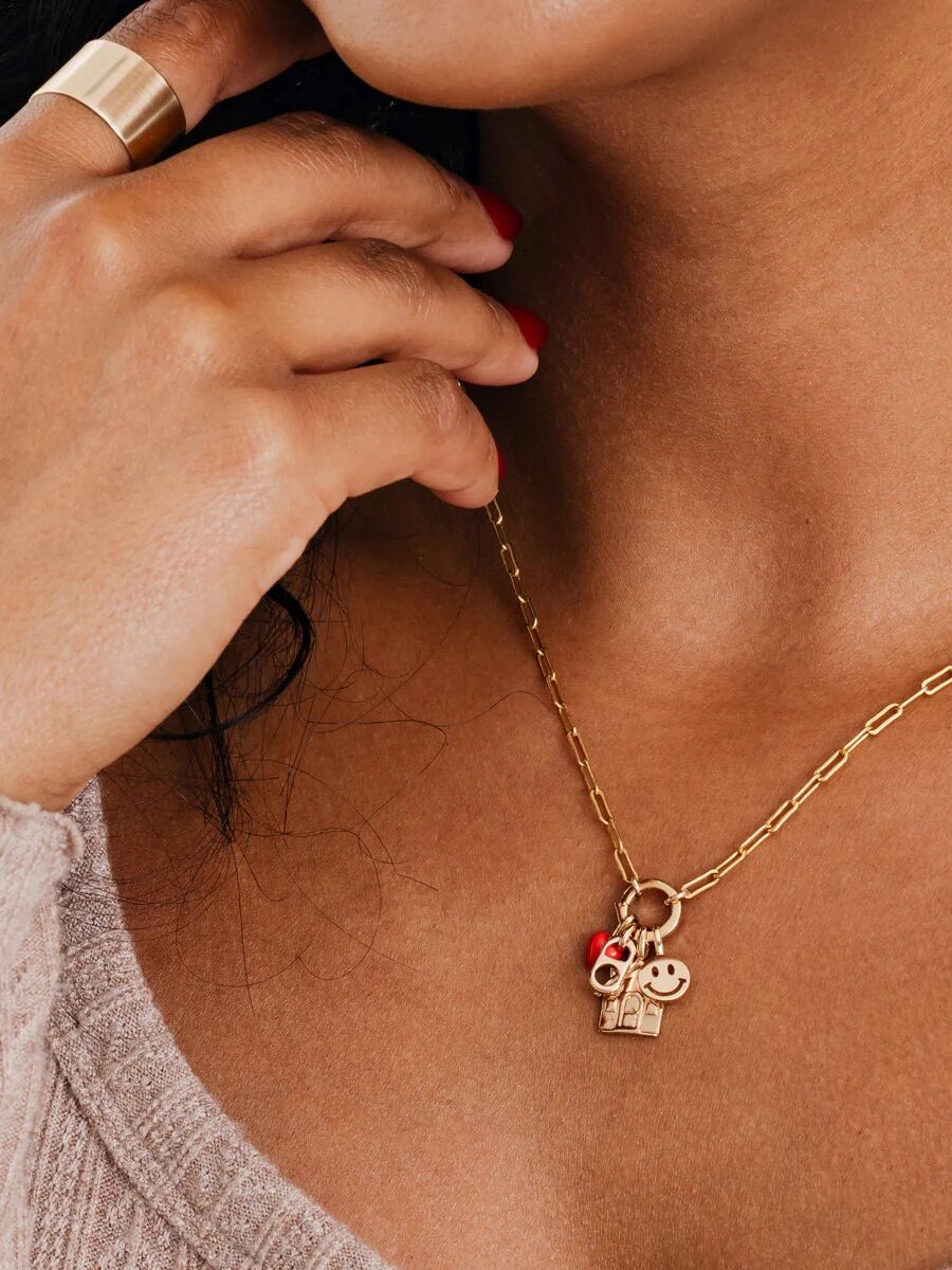 A woman wearing a gold chain necklace with three charms: a smiley face, a gift box, and a red heart. She is holding her chin with fingers adorned with a gold ring.