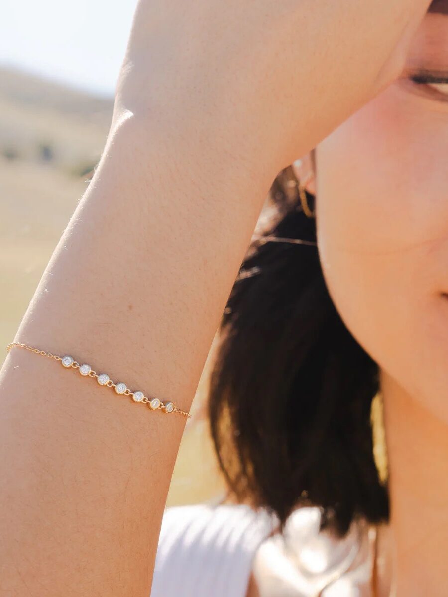 A woman wearing a delicate gold bracelet with small pearls stands outdoors.