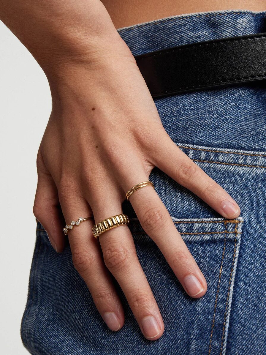 A hand with neatly manicured nails rests on a denim pocket. The fingers wear three gold rings with different designs.