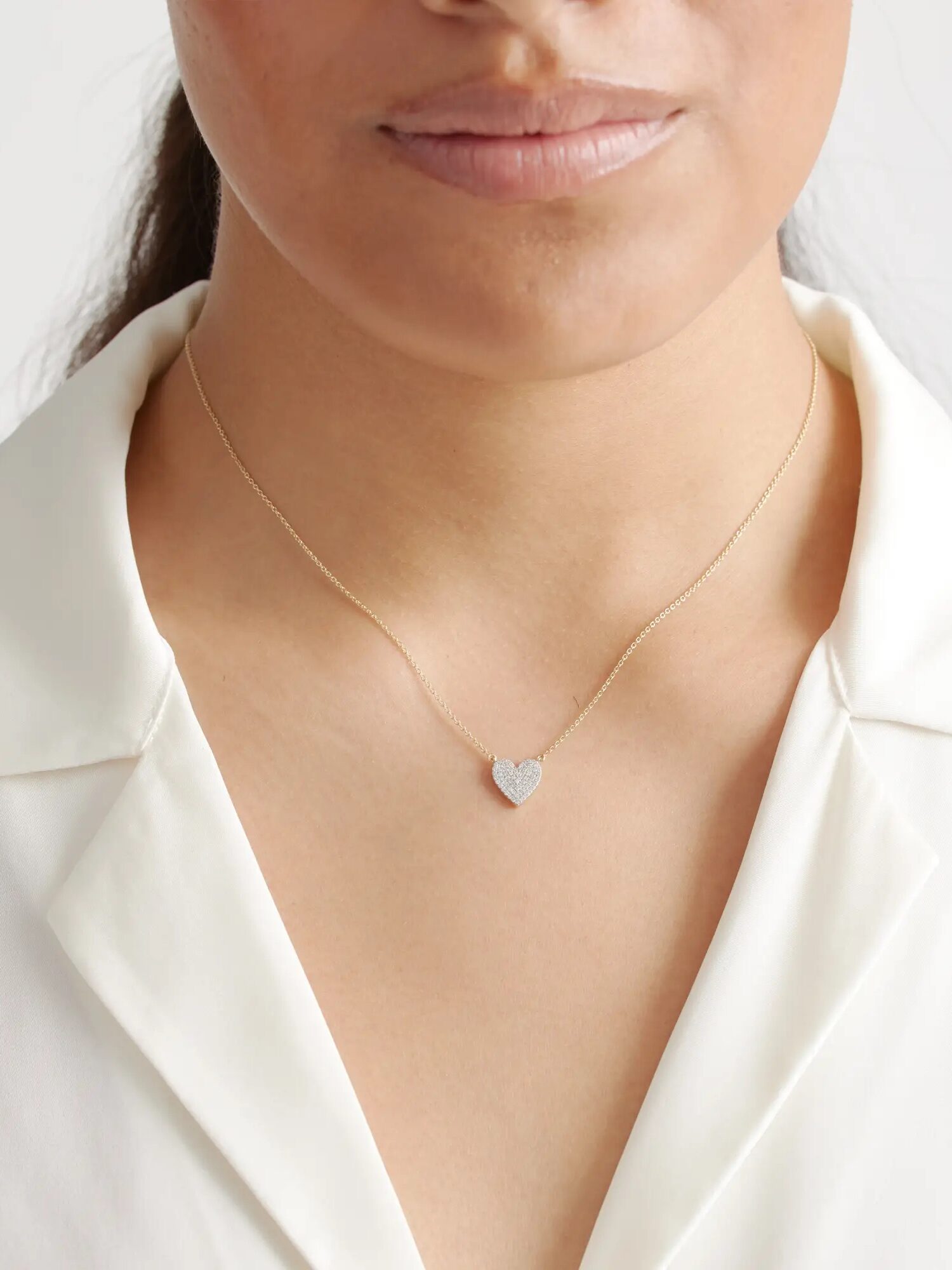 A person wearing a thin chain necklace with a small heart-shaped pendant, dressed in a white button-up shirt.