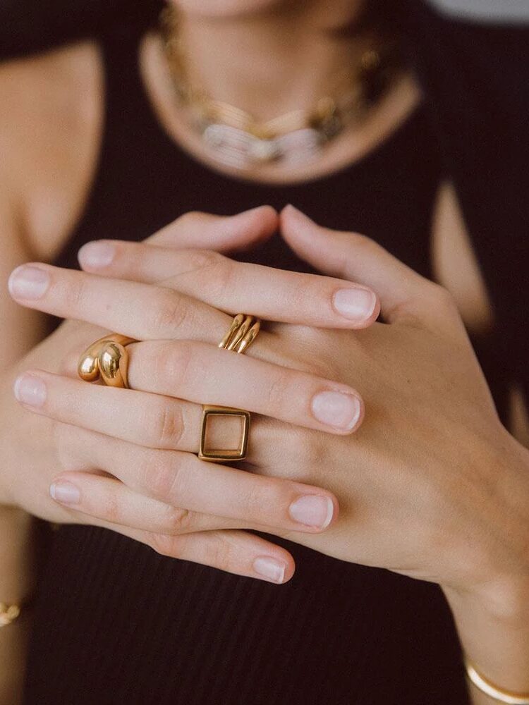 Close-up of a person wearing gold rings and bracelets, with hands clasped over a black top.