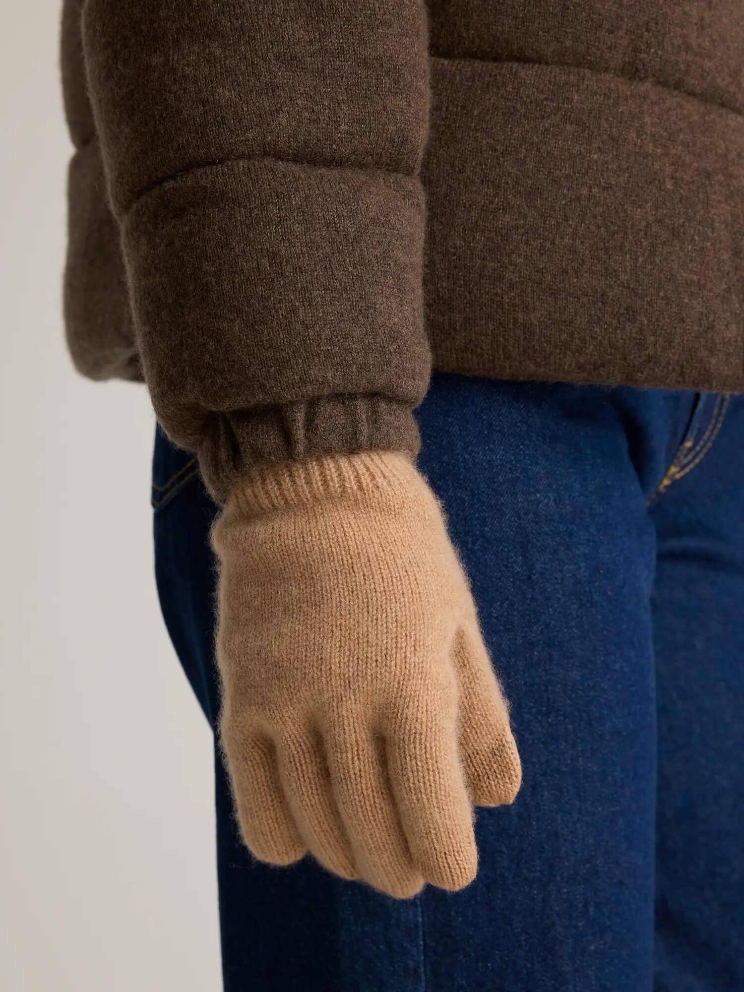 A person wearing a brown coat and blue jeans with a beige glove on their hand.