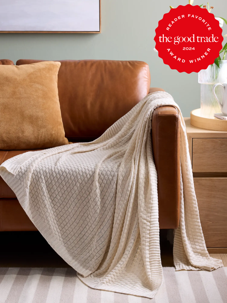 A beige textured throw draped over a brown leather sofa next to a pillow and a wooden side table. A red award badge on the wall reads "Reader Favorite, The Good Trade, 2024 Award Winner.