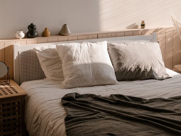 A neatly made bed featuring an organic mattress is adorned with white and gray pillows and a dark gray blanket. The headboard is decorated with small vases, and warm sunlight streams in, creating a cozy atmosphere.