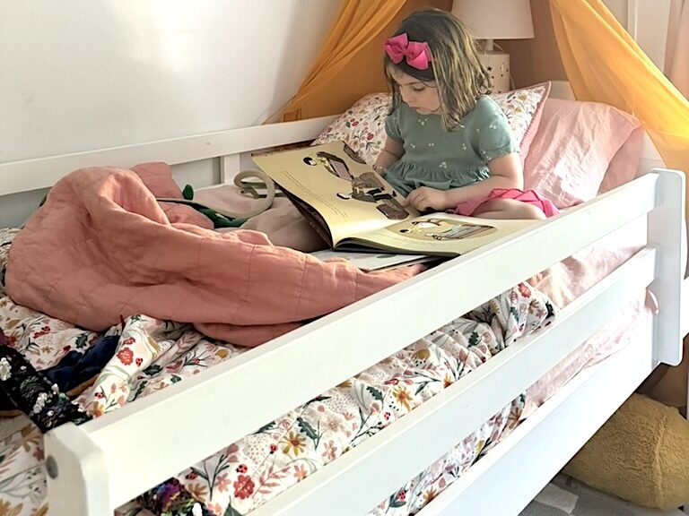 A child in a bed with colorful bedding reads a book under a yellow canopy, nestled comfortably on an organic mattress.