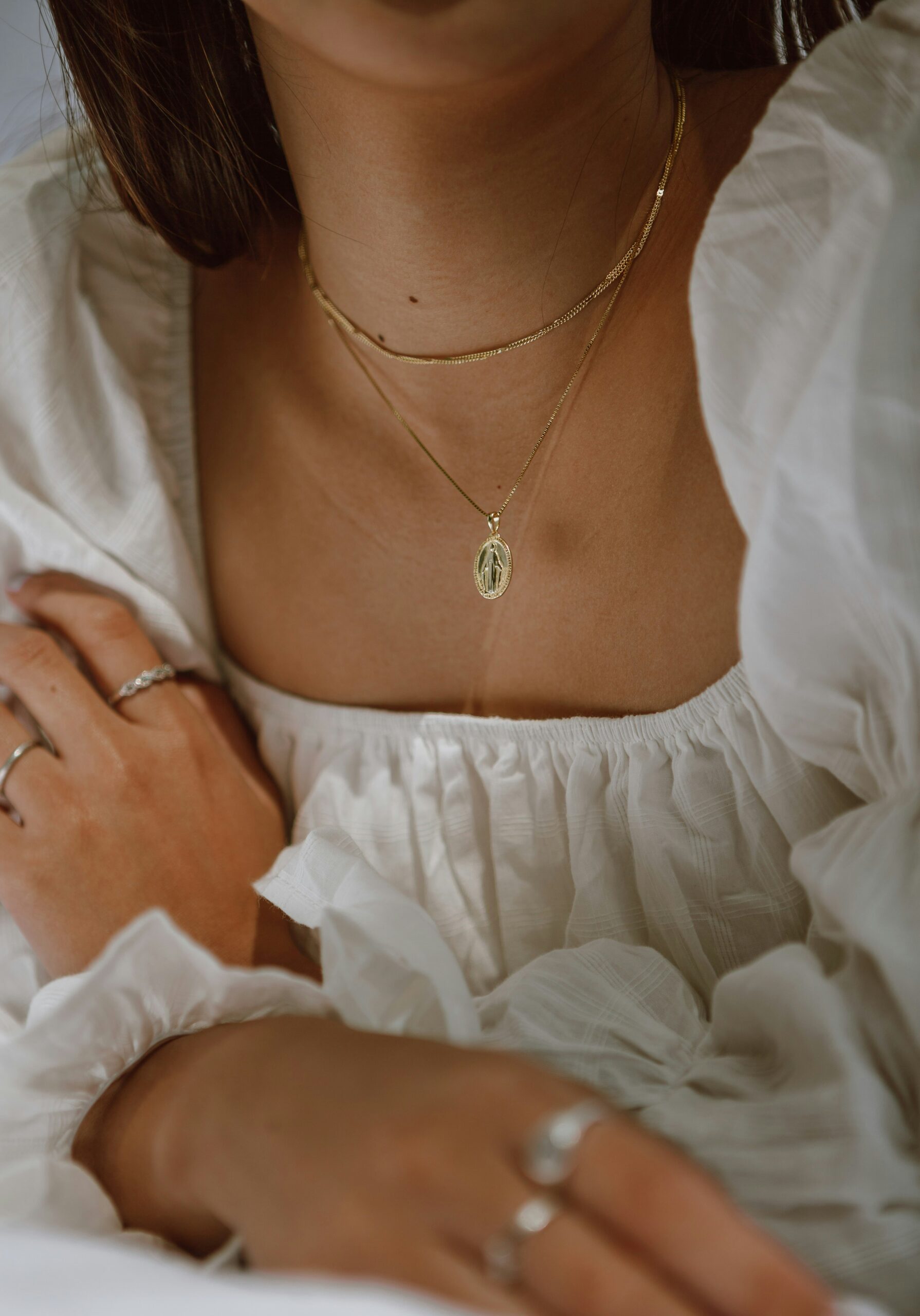 A person wearing a white textured blouse, layered necklaces with a pendant, and silver rings on fingers.