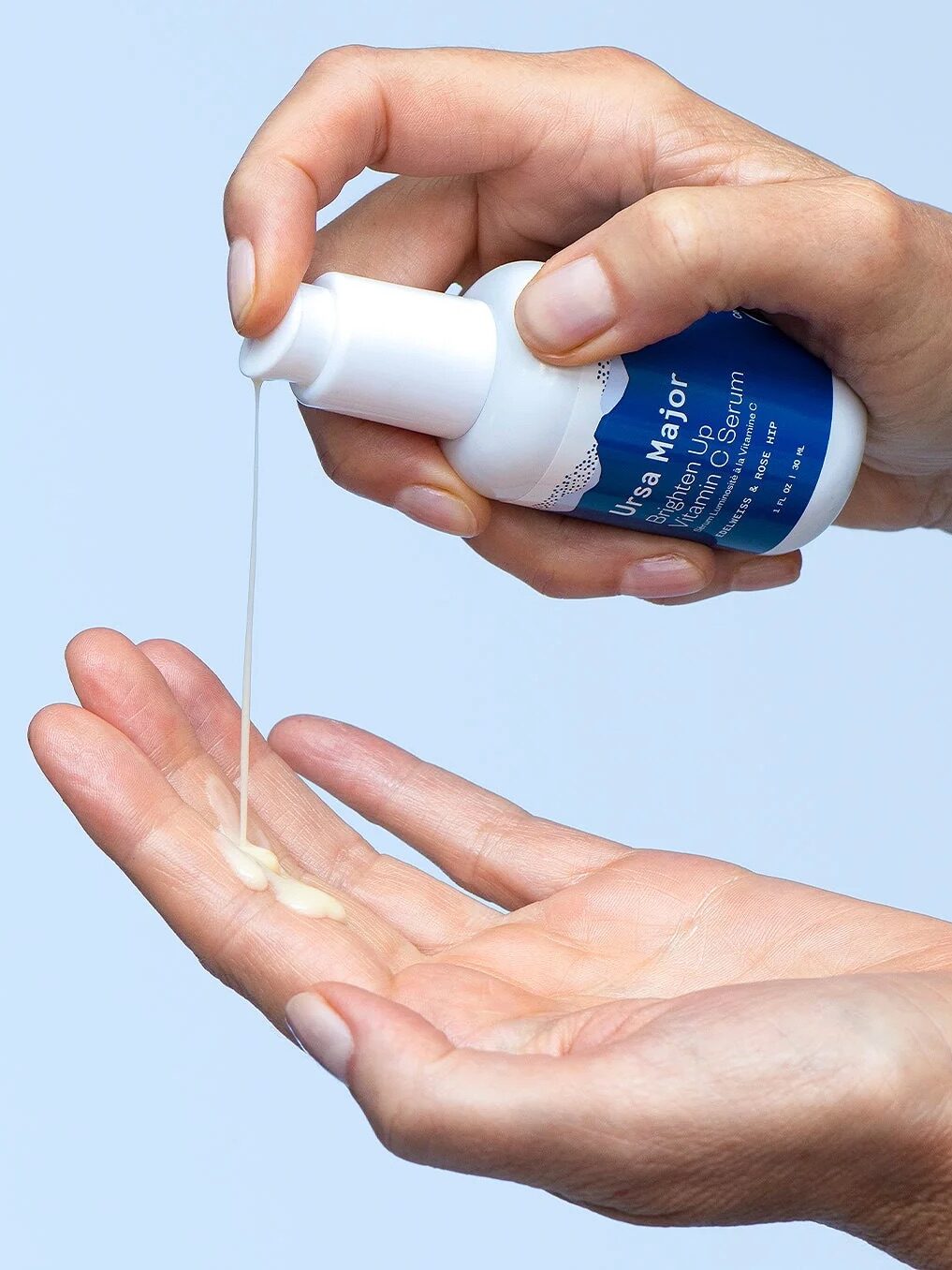 A person dispensing a serum from a white bottle onto their palm against a light blue background.