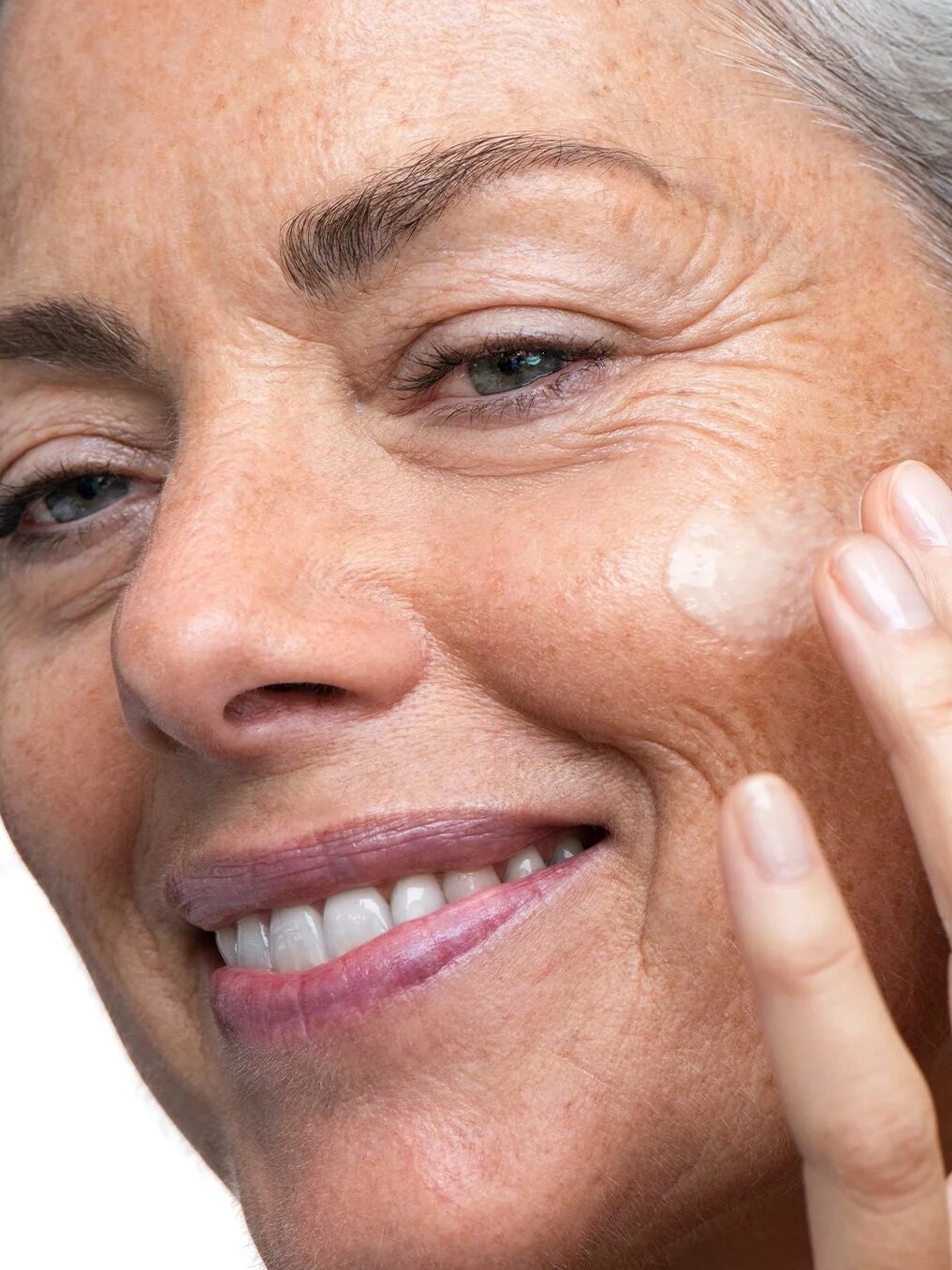 A woman with gray hair applying cream to her cheek, smiling, against a white background.