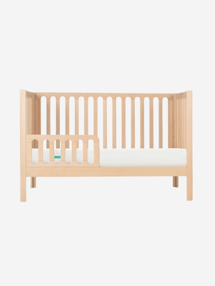 A wooden baby crib with vertical slats and an attached side rail on the left. The mattress is white.