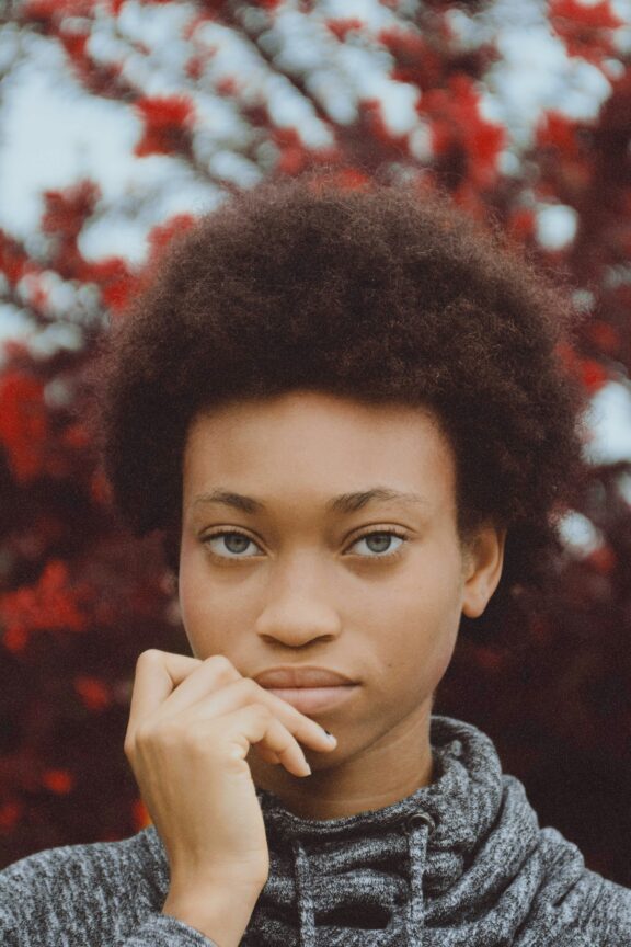 A person with short curly hair, wearing a grey sweater, stands in front of red-leaved trees, looking directly at the camera with one hand touching their chin.