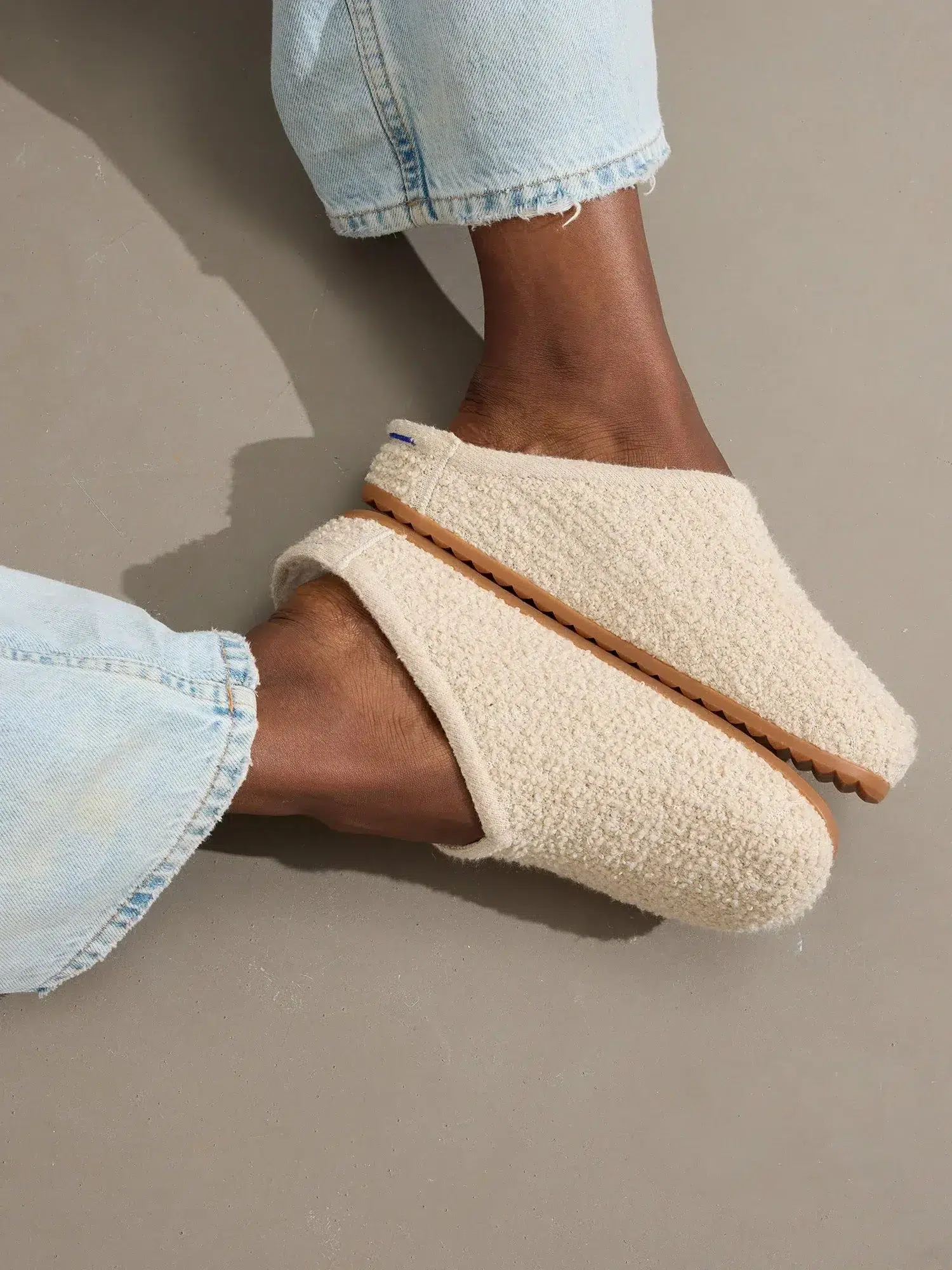 Person wearing light blue jeans and beige fuzzy slippers sitting on a gray surface.