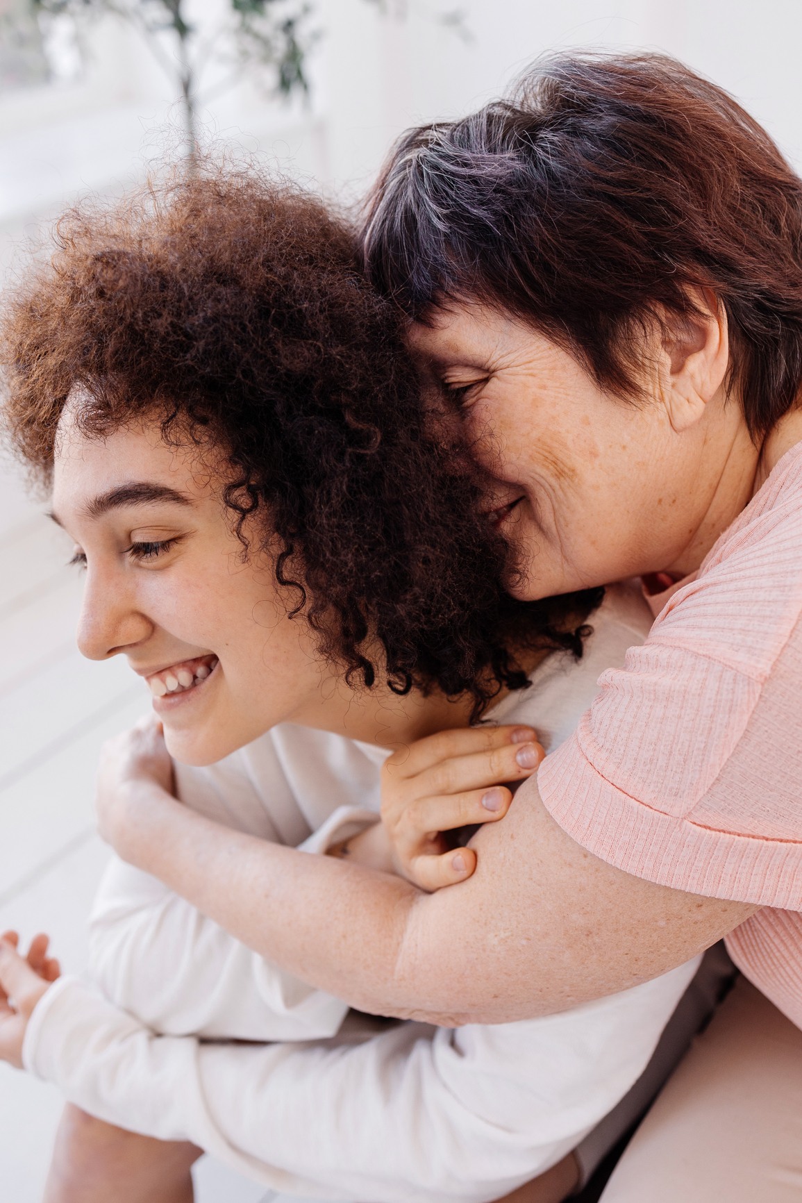 Two people sitting closely; one with curly hair smiling, receiving a side hug from another with short hair, who is also smiling.