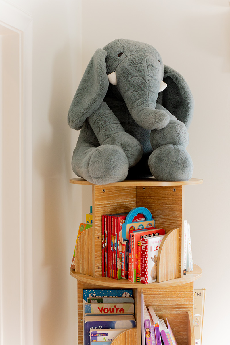 A large plush elephant sits atop a wooden corner bookshelf filled with children's books.