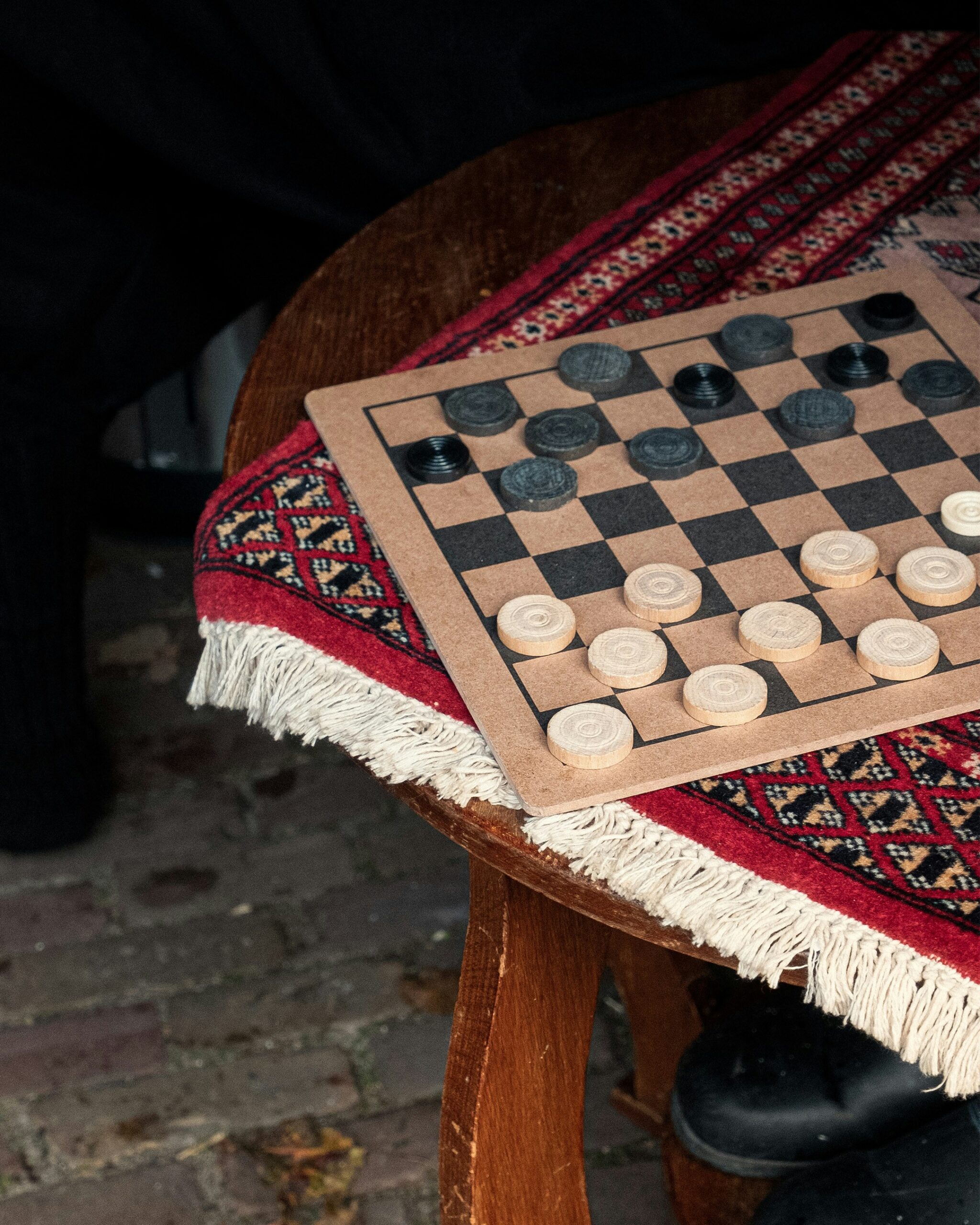 A checkers board on a wooden table with a fringed red patterned cloth underneath. The game is in progress with black and white pieces positioned.