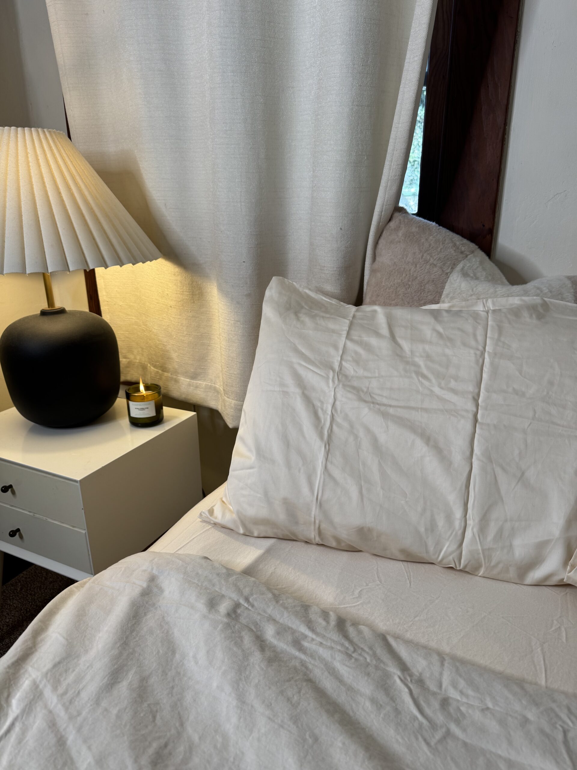 A cozy bedroom scene with a neatly made bed featuring cream linens, a pillow, a small white bedside table, a lit candle, and a lamp with a pleated shade.