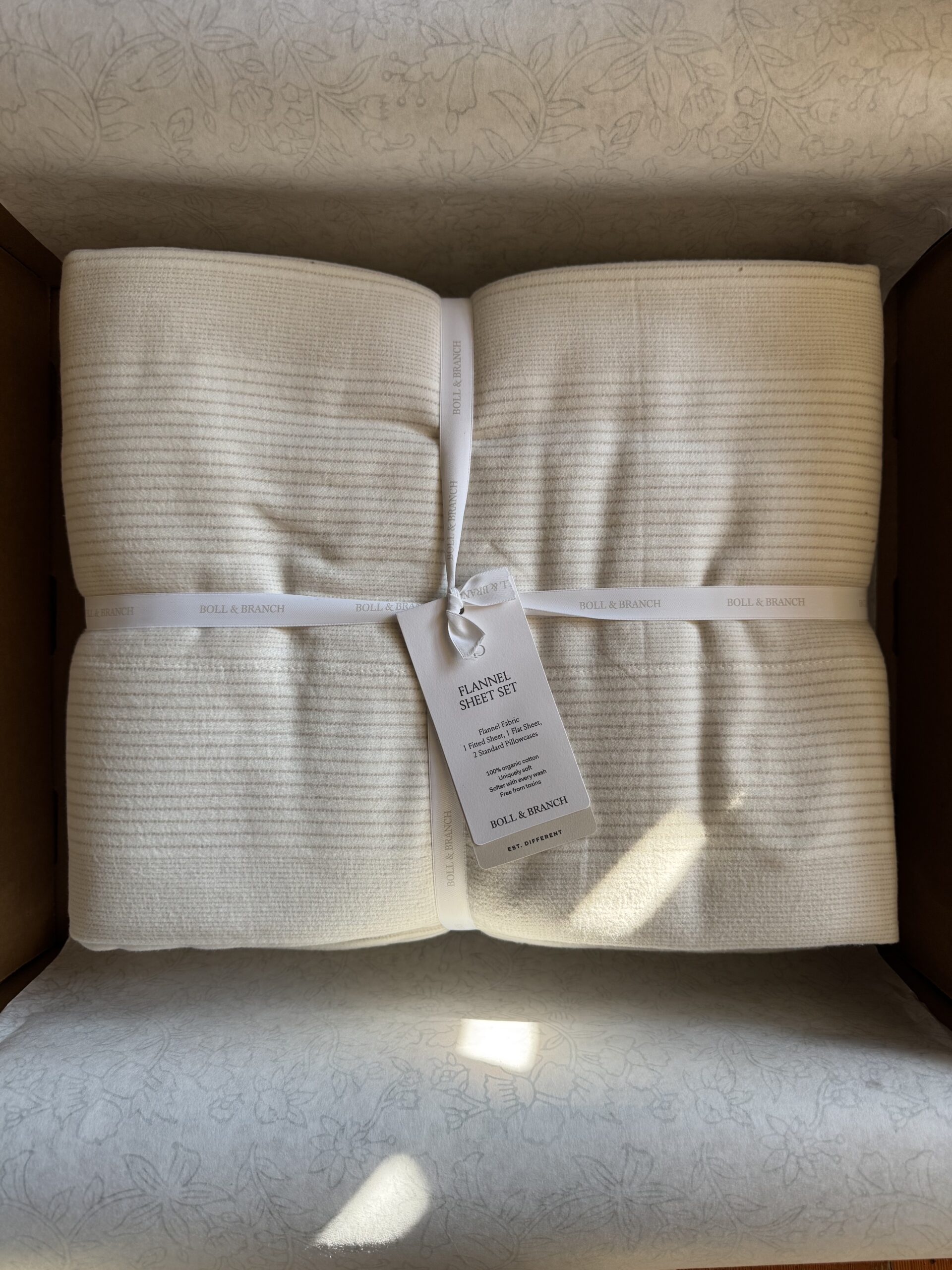 A neatly folded, cream-colored flannel sheet set tied with a white ribbon, placed in a cardboard box. A tag with product details is attached.