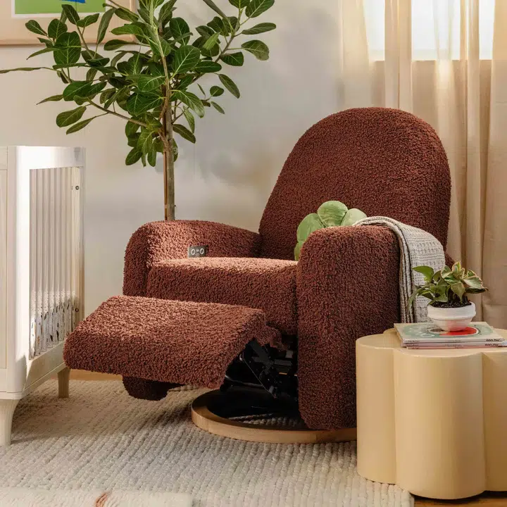 A brown textured recliner next to a small table with books and plants. A potted tree is in the background, near a window with beige curtains.