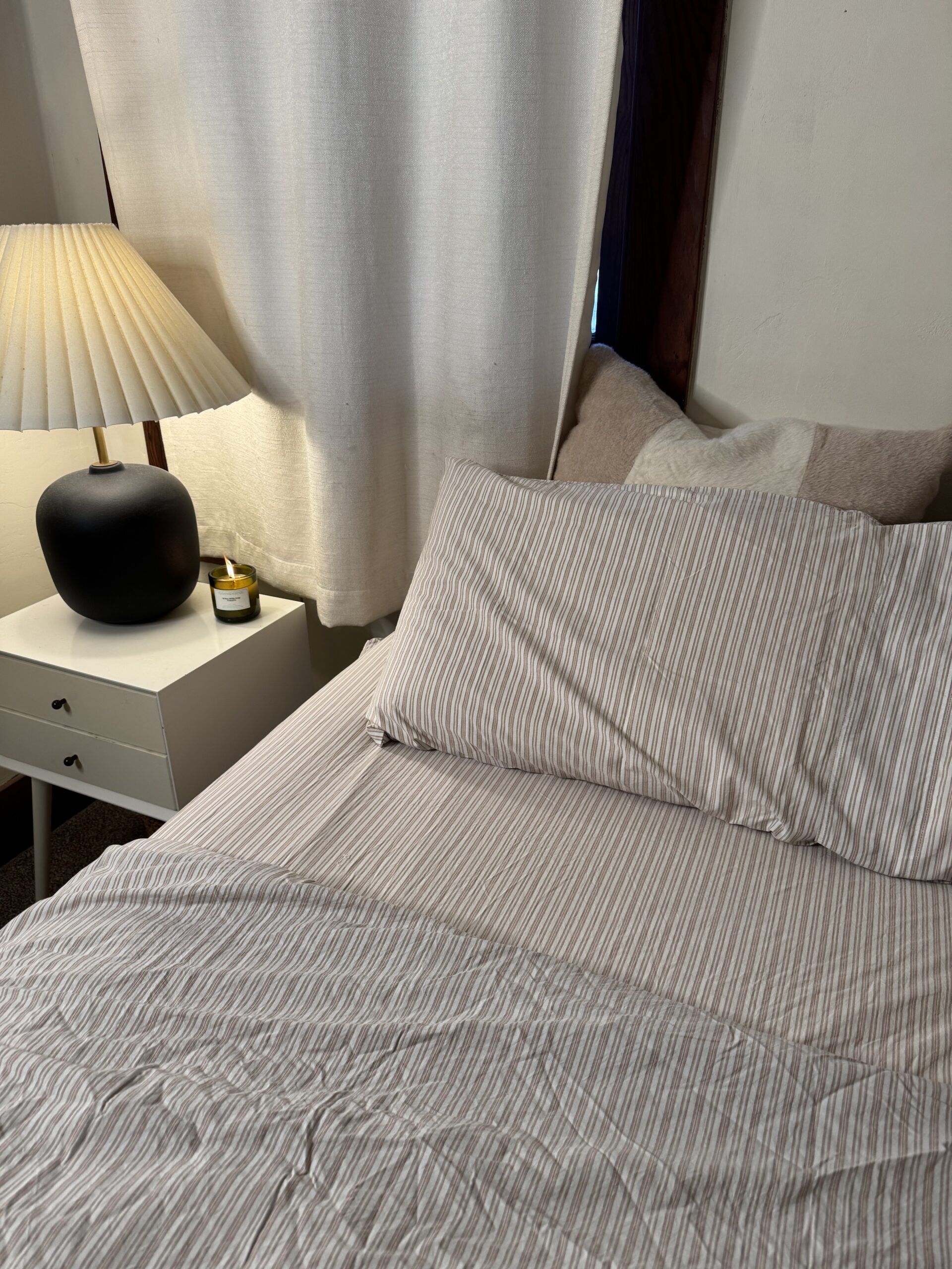 A neatly made bed with striped sheets is next to a nightstand holding a lamp and a small candle. White curtains hang by the window.