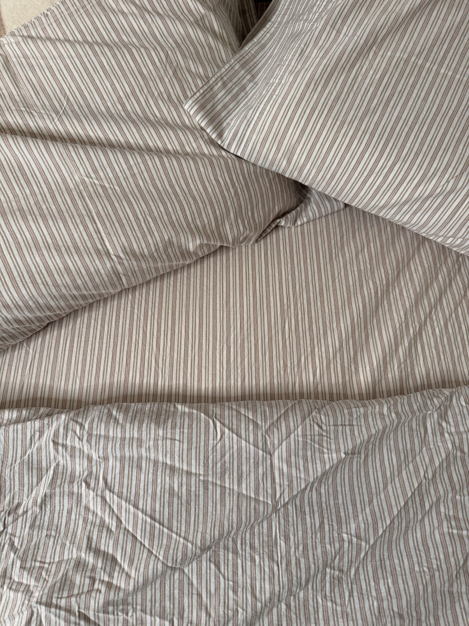 A bed with beige and white striped sheets and pillowcases, showing a slightly wrinkled appearance.