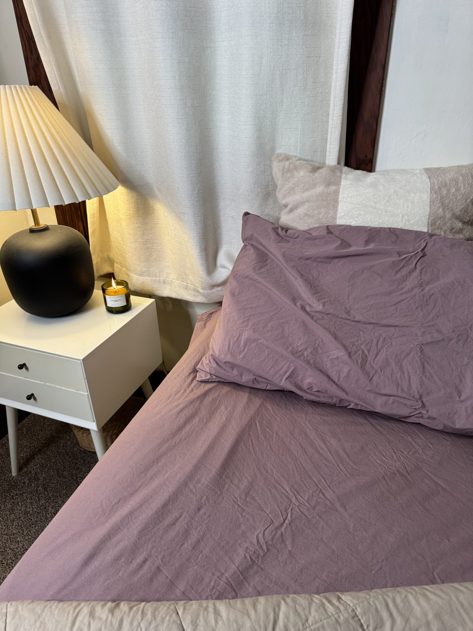 A neatly made bed with purple sheets and a pillow is next to a white nightstand. A lit candle and a lamp with a pleated shade are on the nightstand. White curtains hang nearby.