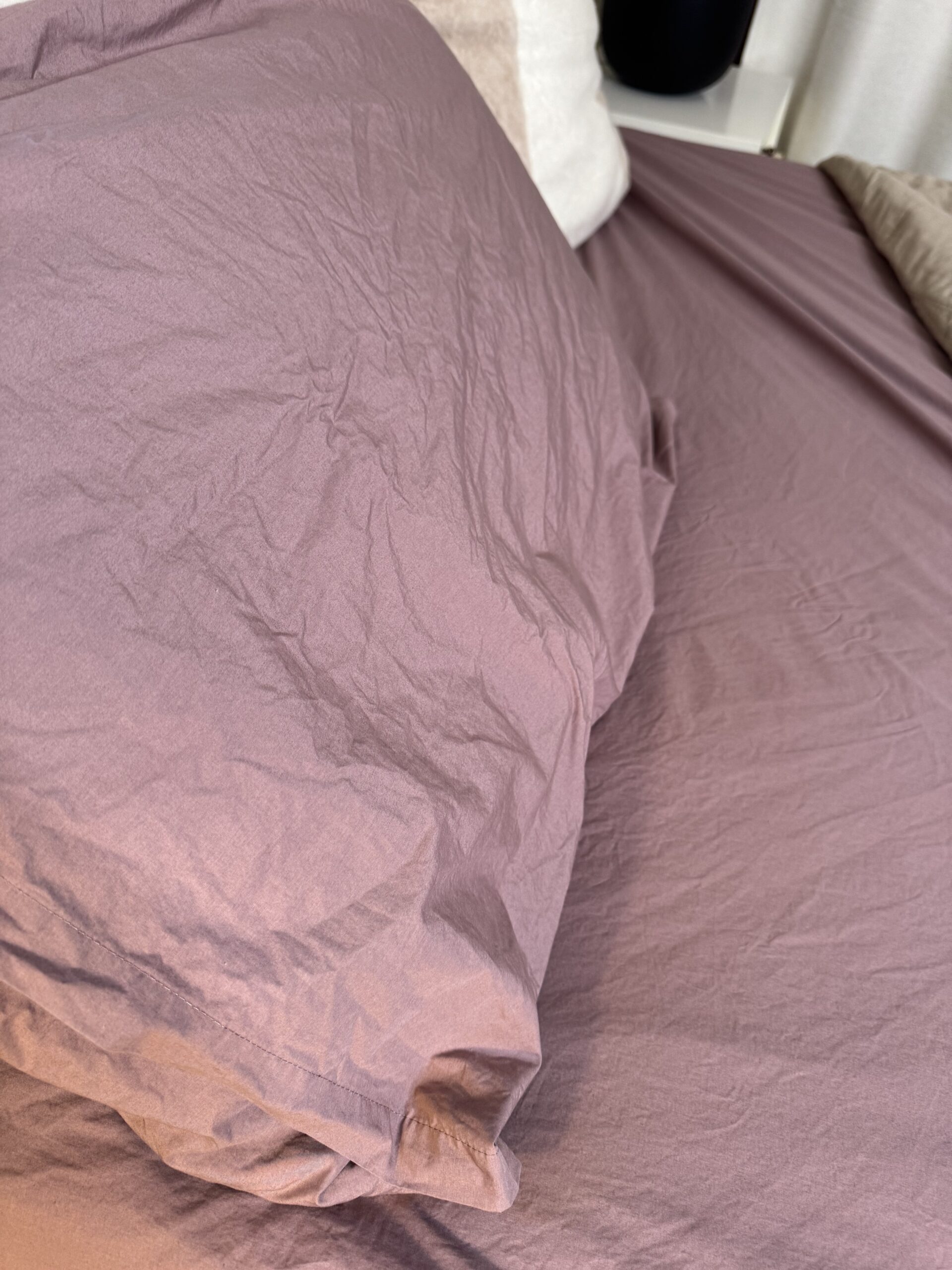 A bed with a mauve pillow and matching crumpled sheet.