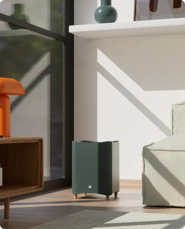 A modern, black air purifier is placed on a light wooden floor next to a green armchair and under a white shelf with decorative items. Sunlight streams through a window.