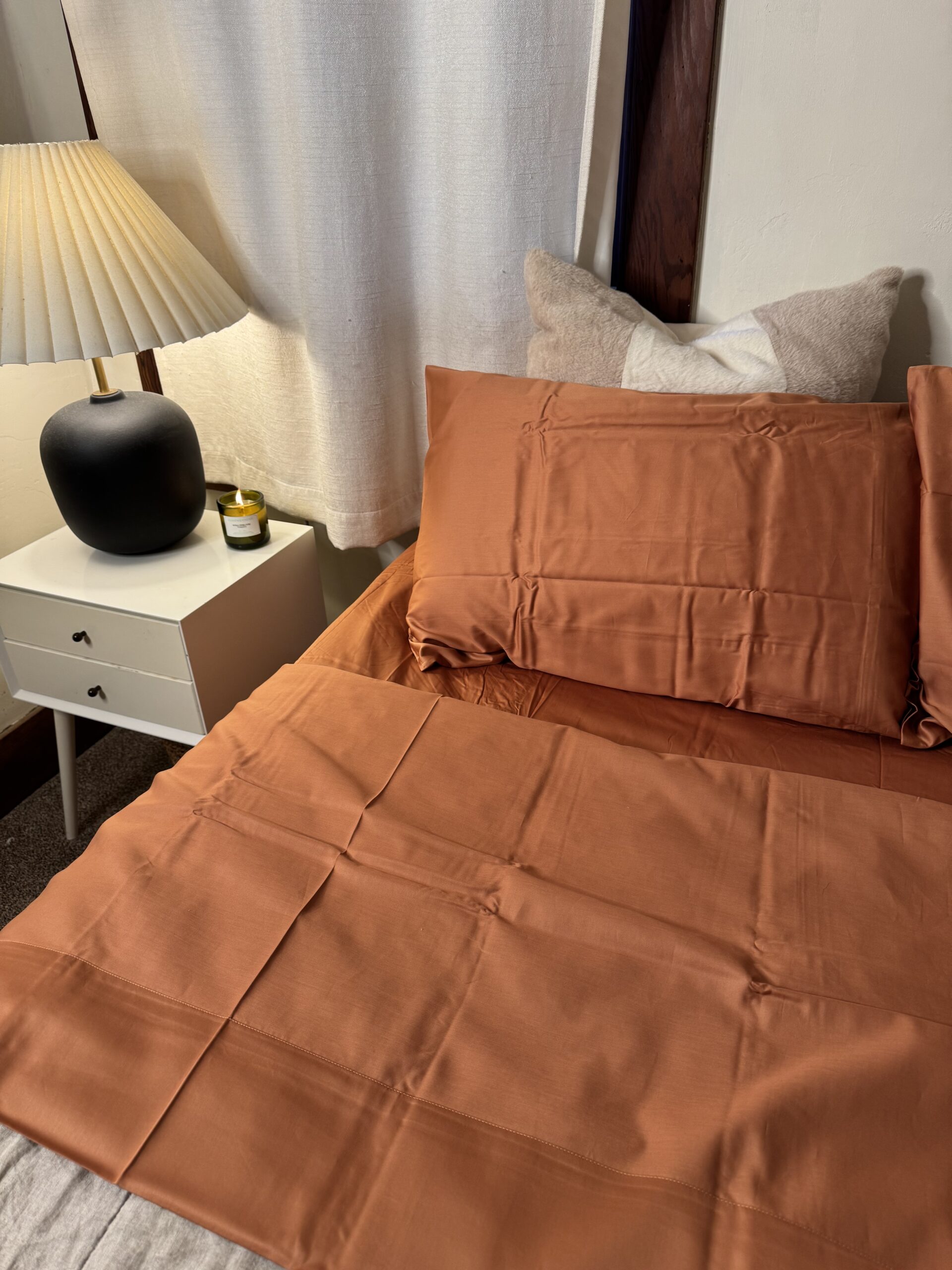 A bed with rust-colored sheets and matching pillowcases, next to a white nightstand with a black lamp and a candle. A white curtain hangs in the background.