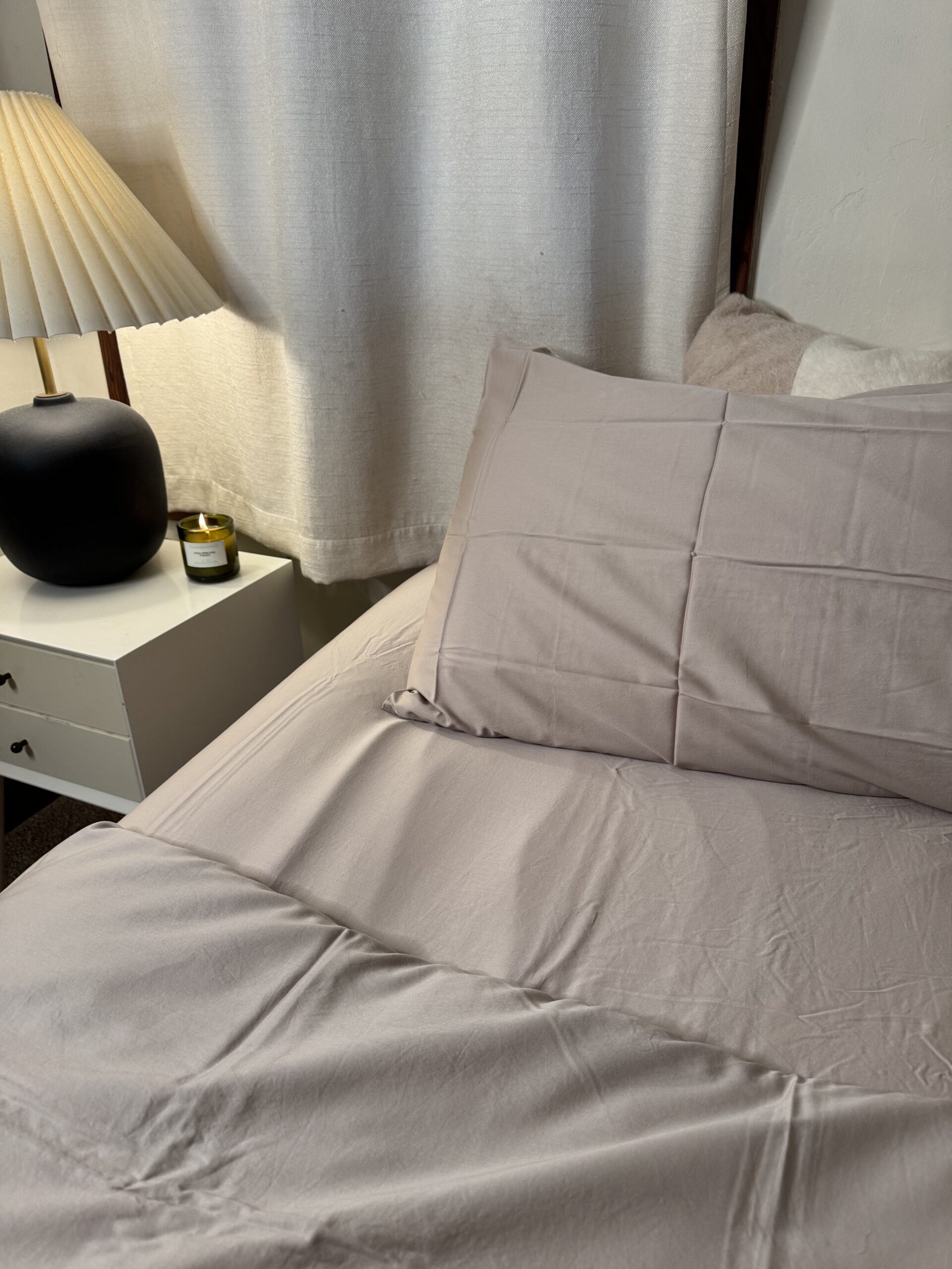 A neatly made bed with beige sheets and pillows next to a nightstand. A lamp, black vase, and candle sit on the nightstand. White curtains hang in the background.