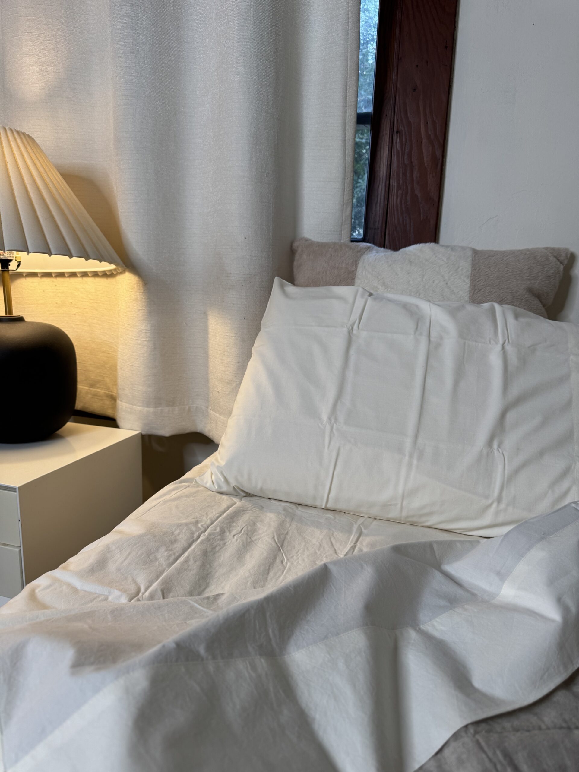 A neatly made bed with a white pillow and bedspread, beside a lit table lamp on a nightstand, in front of a curtain-covered window.