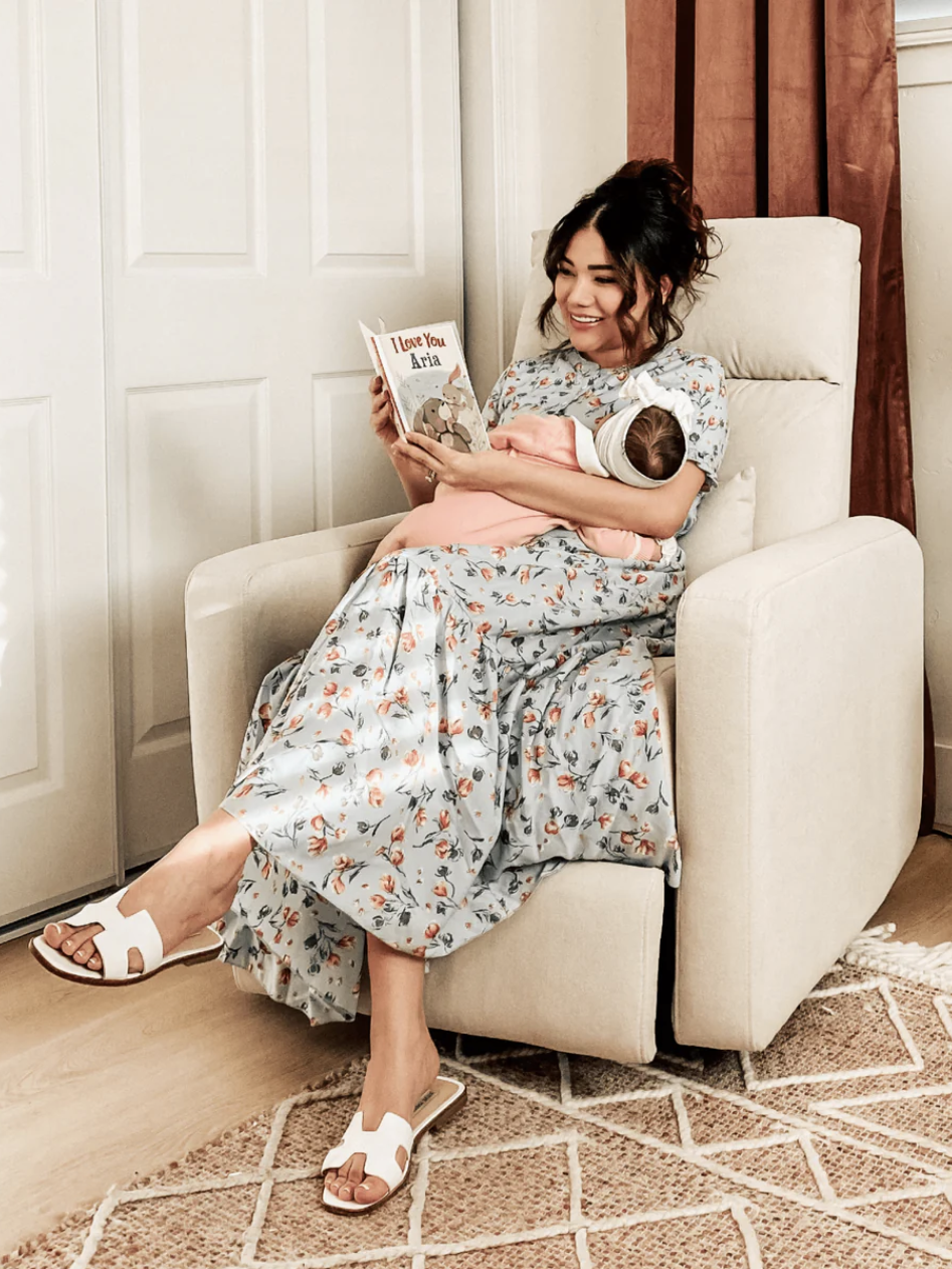 A woman in a floral dress sits in an armchair reading a book titled "I Love You, Alex" to a baby resting on her lap in a cozy room.