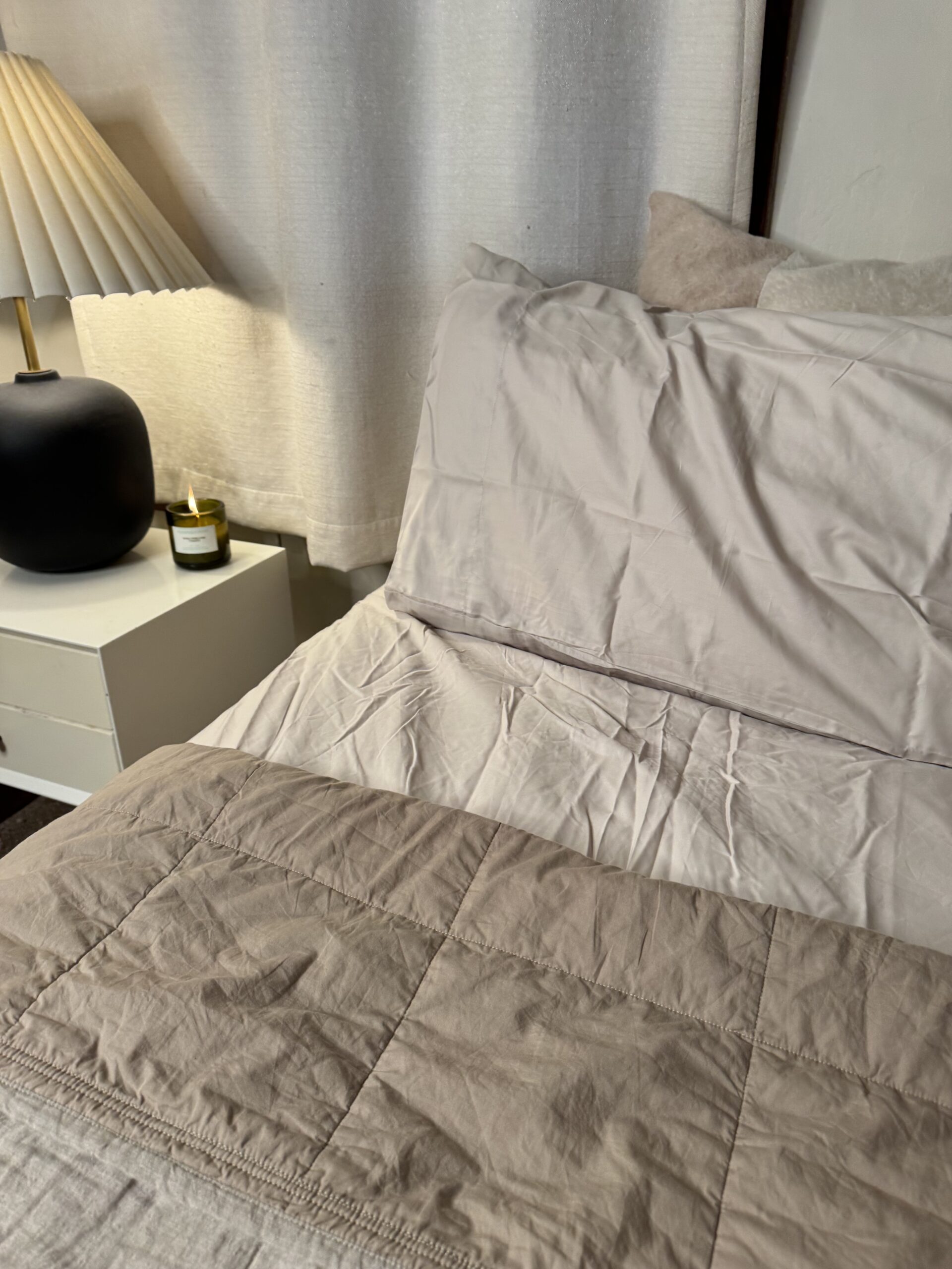 Unmade bed with white sheets, a beige duvet, and pillows. A bedside table holds a black lamp and a candle, with a curtain in the background.